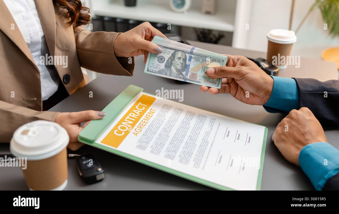 Un uomo d'affari e un cliente stanno esaminando e firmando un contratto di vendita di auto presso una scrivania, con modellini di auto di piccole dimensioni, una tazza di caffè e documenti intorno a loro, Foto Stock