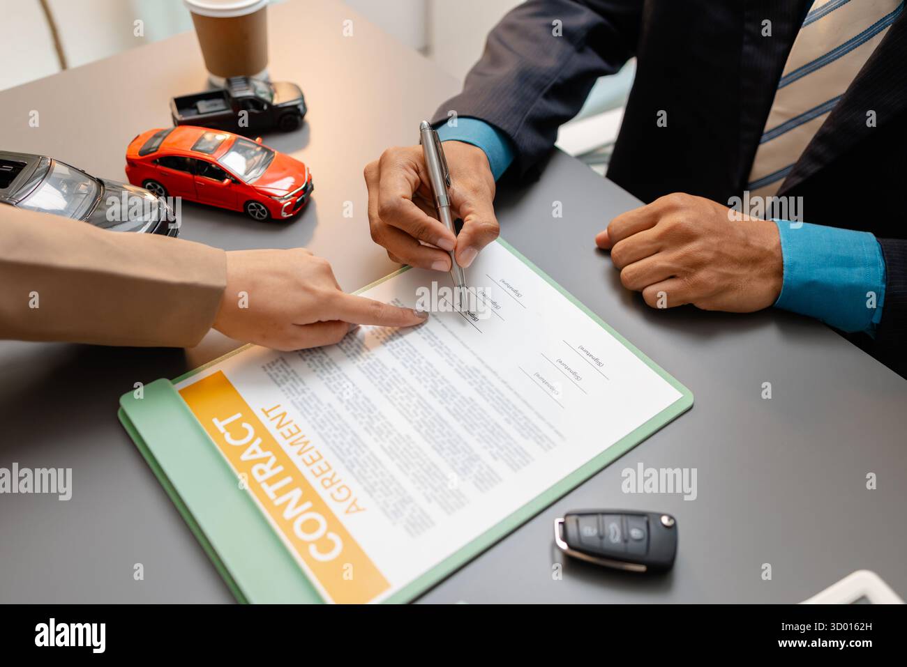 Un uomo d'affari e un cliente stanno esaminando e firmando un contratto di vendita di auto presso una scrivania, con modellini di auto di piccole dimensioni, una tazza di caffè e documenti intorno a loro, Foto Stock