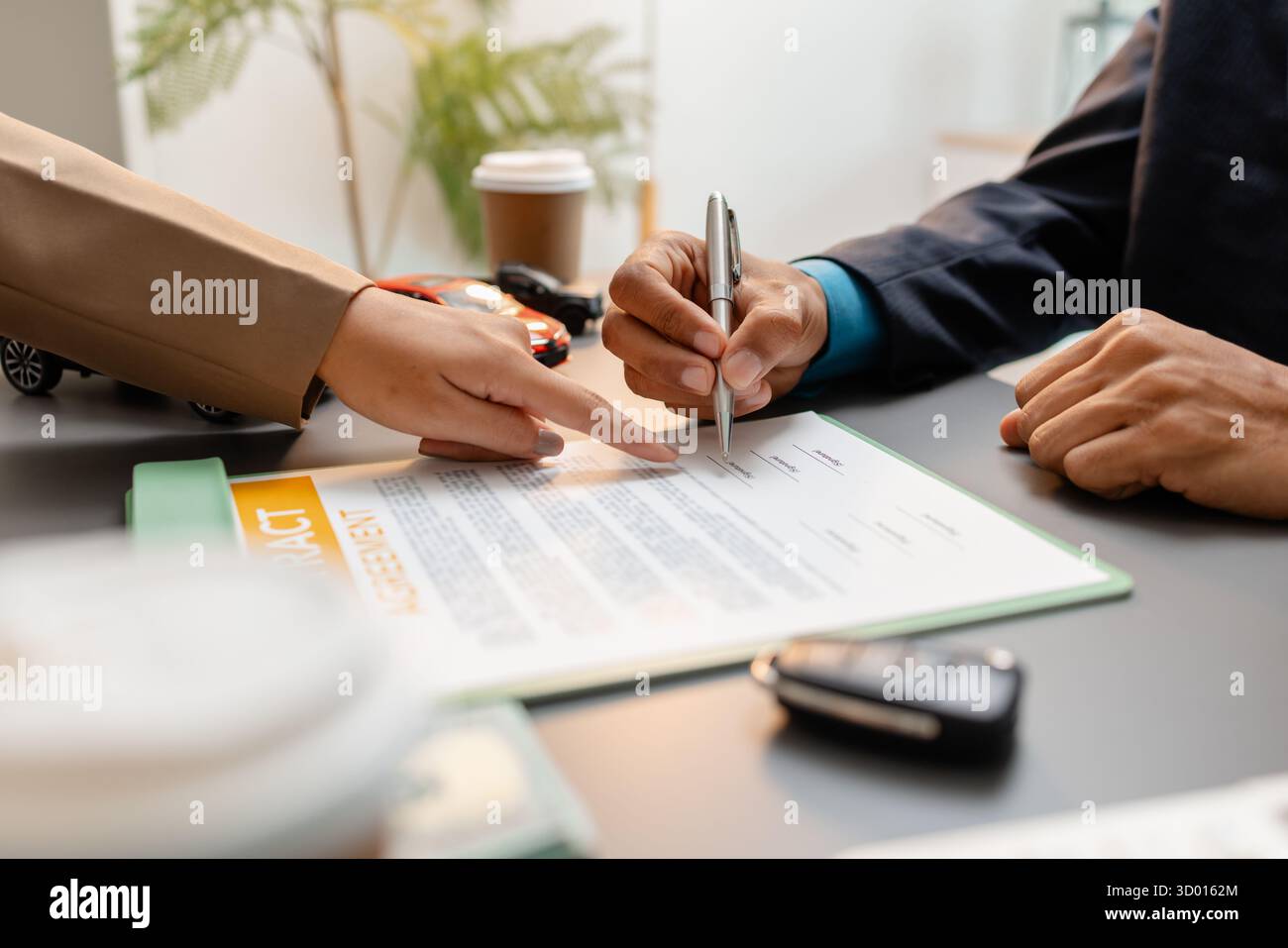 Un uomo d'affari e un cliente stanno esaminando e firmando un contratto di vendita di auto presso una scrivania, con modellini di auto di piccole dimensioni, una tazza di caffè e documenti intorno a loro, Foto Stock