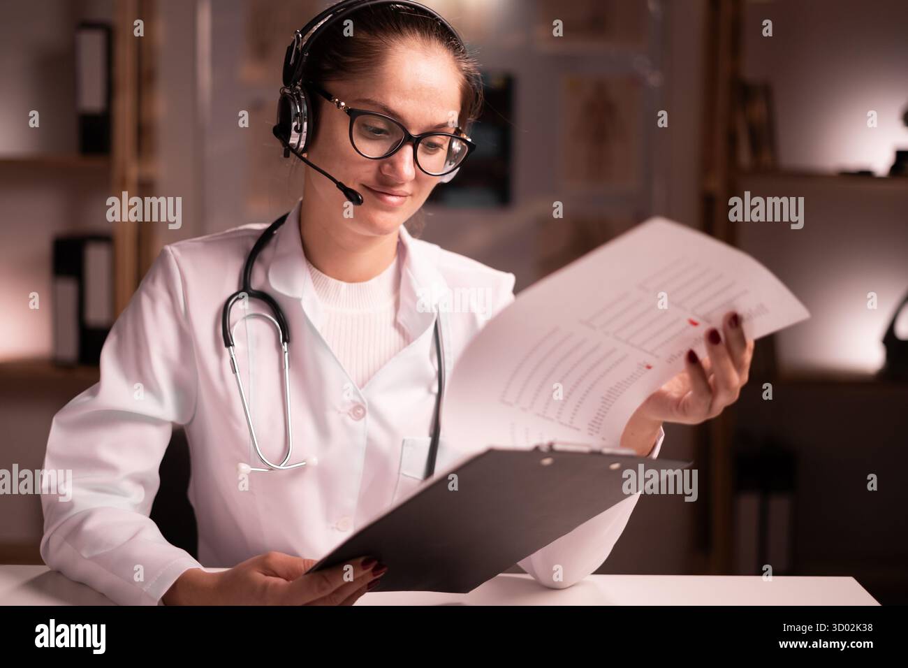 medico donna in camice bianco con cuffia e appunti consulenza paziente tramite videochiamata, conferenza sanitaria online, lettura elenco dei farmaci Foto Stock