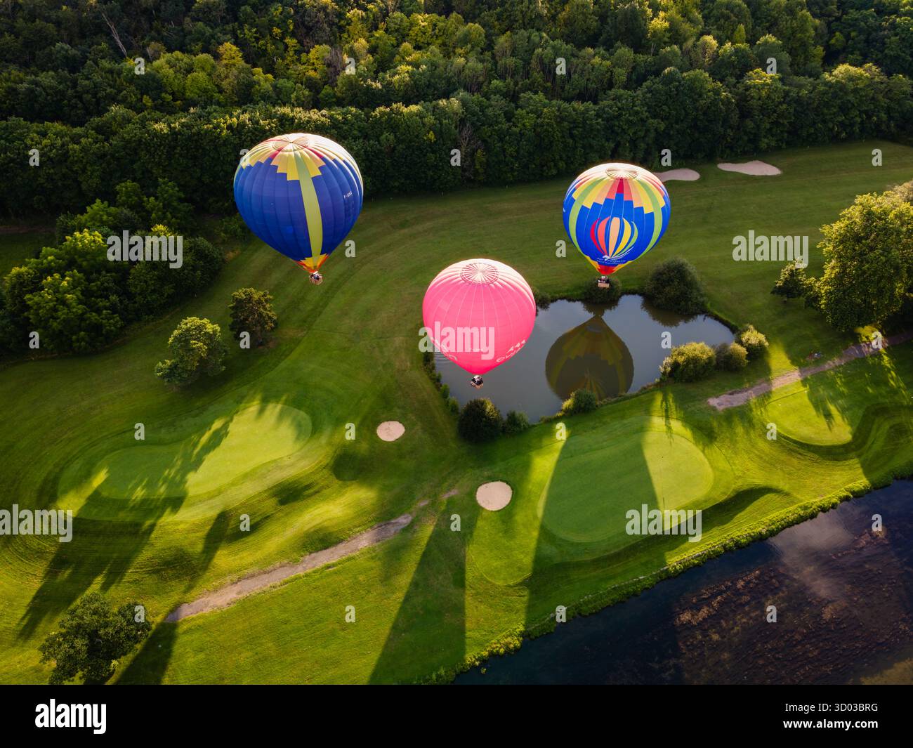 Ripresa aerea dall'alto verso il basso di tre vivaci mongolfiere che salgono da un parco verde durante l'ora d'oro, catturando motivi colorati Foto Stock