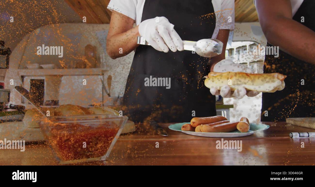 Colleghi che preparano salsicce che riempiono panini hot dog nella cucina del caffè, con pinze in metallo e ciotola per peperoncino Foto Stock