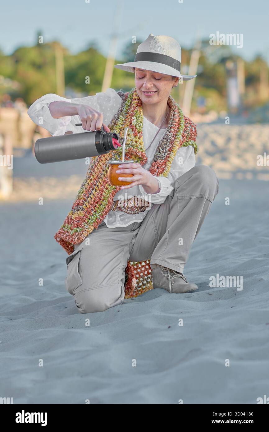 Una donna matura prepara il suo compagno su una spiaggia uruguaiana, circondata da luce calda e sabbia. Vestita in stile bohémien, incarna un momento intimo Foto Stock