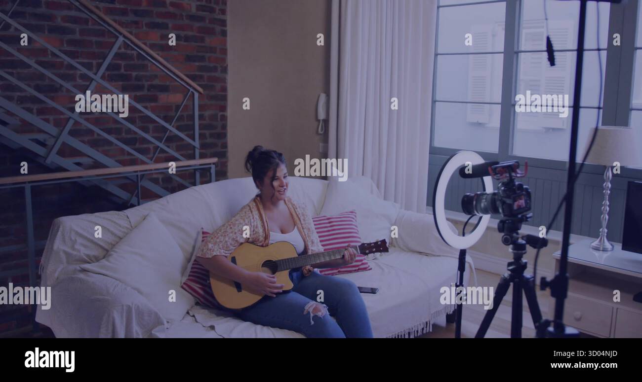 Donna che suona jeans strappati sul divano del soggiorno, con chitarra acustica e configurazione della fotocamera Foto Stock