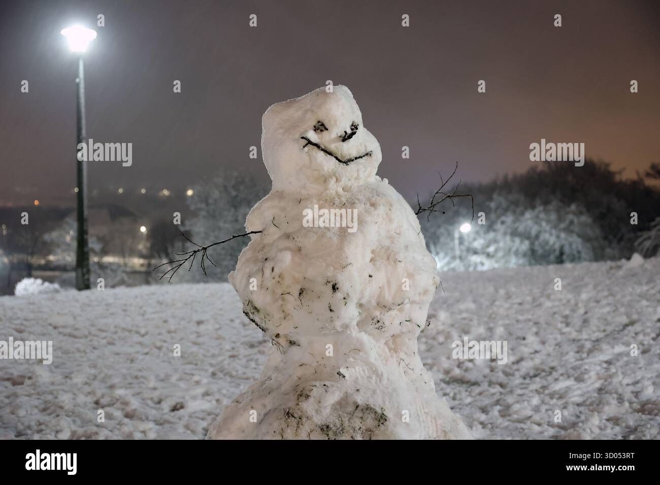 Pupazzo di neve fatto a mano con le braccia del bastone e il viso in piedi su una collina innevata di notte sotto il bagliore dei lampioni Foto Stock
