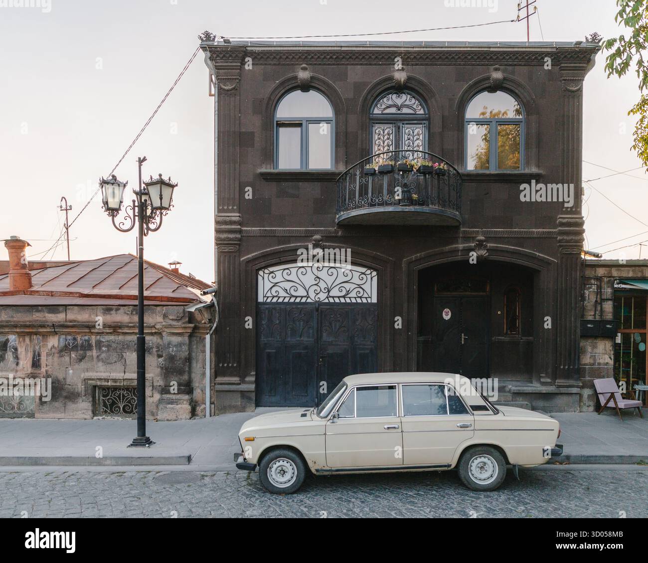 Vista di un'auto color crema d'epoca parcheggiata davanti a un edificio in pietra scura con finestre ad arco e un balcone in ferro battuto, Gyumri, provincia di Shirak, Armenia. Foto Stock