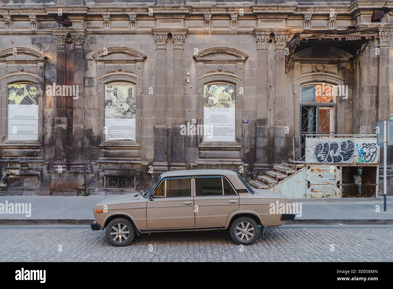 Vista di una vecchia auto parcheggiata di fronte a un edificio intempestivo con poster sbiaditi e graffiti, che evocano un senso di storia e degrado urbano, Gyumri, provincia di Shirak, Armenia. Foto Stock