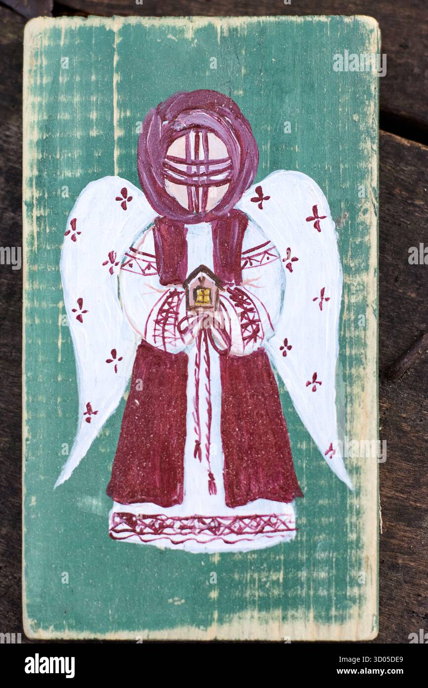 Un angelo di legno fatto a mano con ali e un mantello rosso regge una casa, raffigurata su uno sfondo verde, creando un ornamento natalizio di arte popolare. Foto Stock