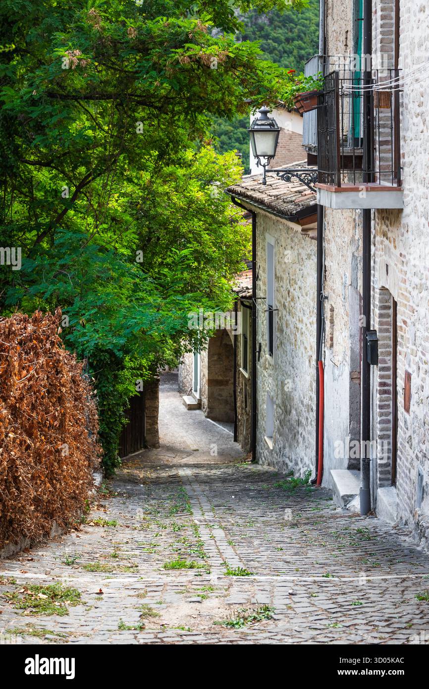 La vista del sentiero in pietra abbellito dal sole si snoda attraverso vecchi edifici, abbracciati da una vegetazione lussureggiante in un villaggio italiano senza tempo, Anversa degli Abruzzi, Abruzzo, Italia. Foto Stock