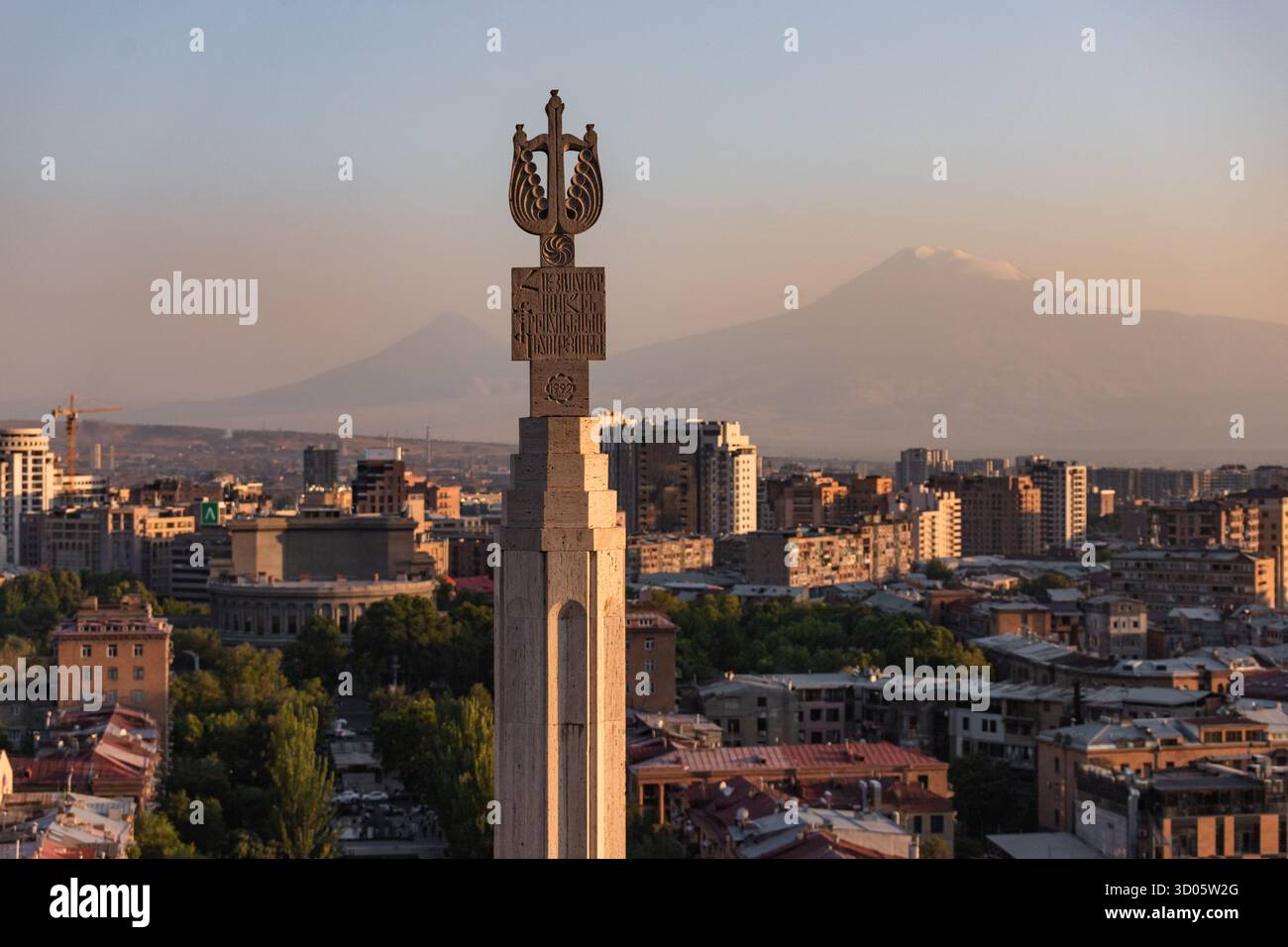 Vista del pilastro in pietra scolpita che sorge sopra un vibrante paesaggio urbano mentre il sole dipinge il cielo in tonalità calde, con maestose montagne in lontananza, Yerevan, Yer Foto Stock