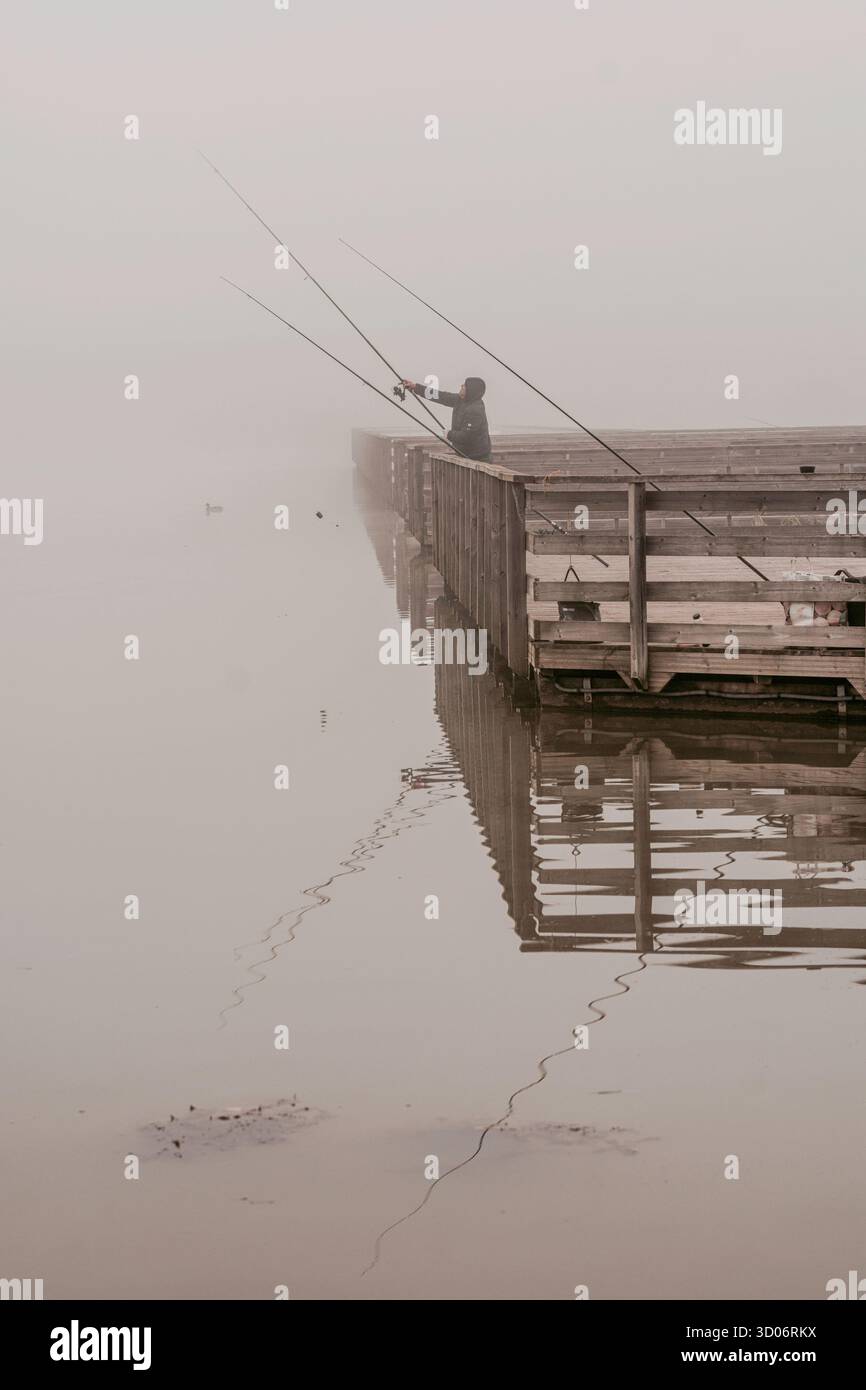 Bacau, Romania, 18 ottobre 2025: Persona non identificabile che pesca da un molo di legno in fitta nebbia. Foto Stock