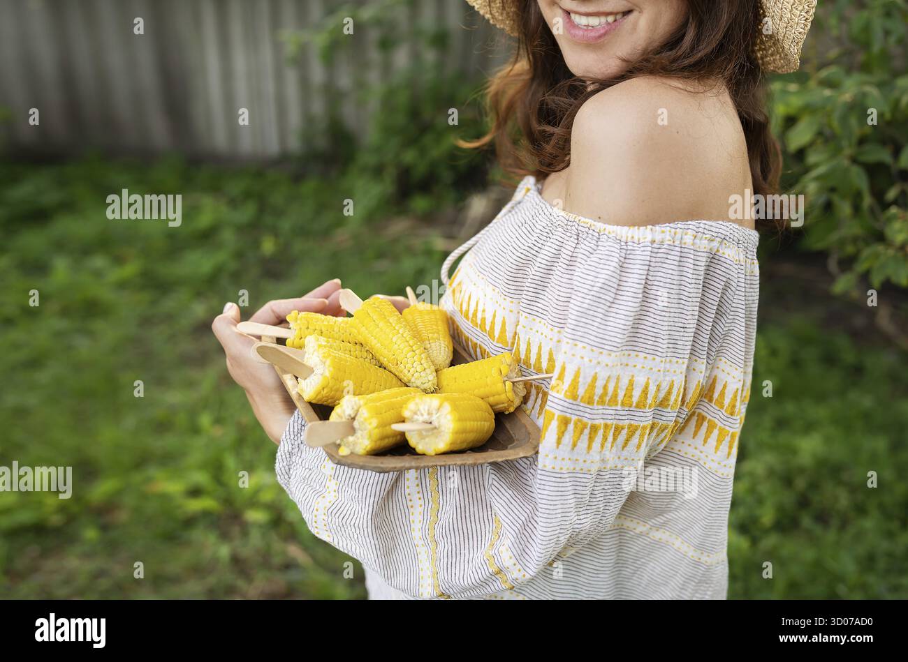 La ragazza sorride in un cappello di paglia e tiene in mano un piatto con mais bollito. Il concetto di attività ricreative all'aperto, barbecue Foto Stock