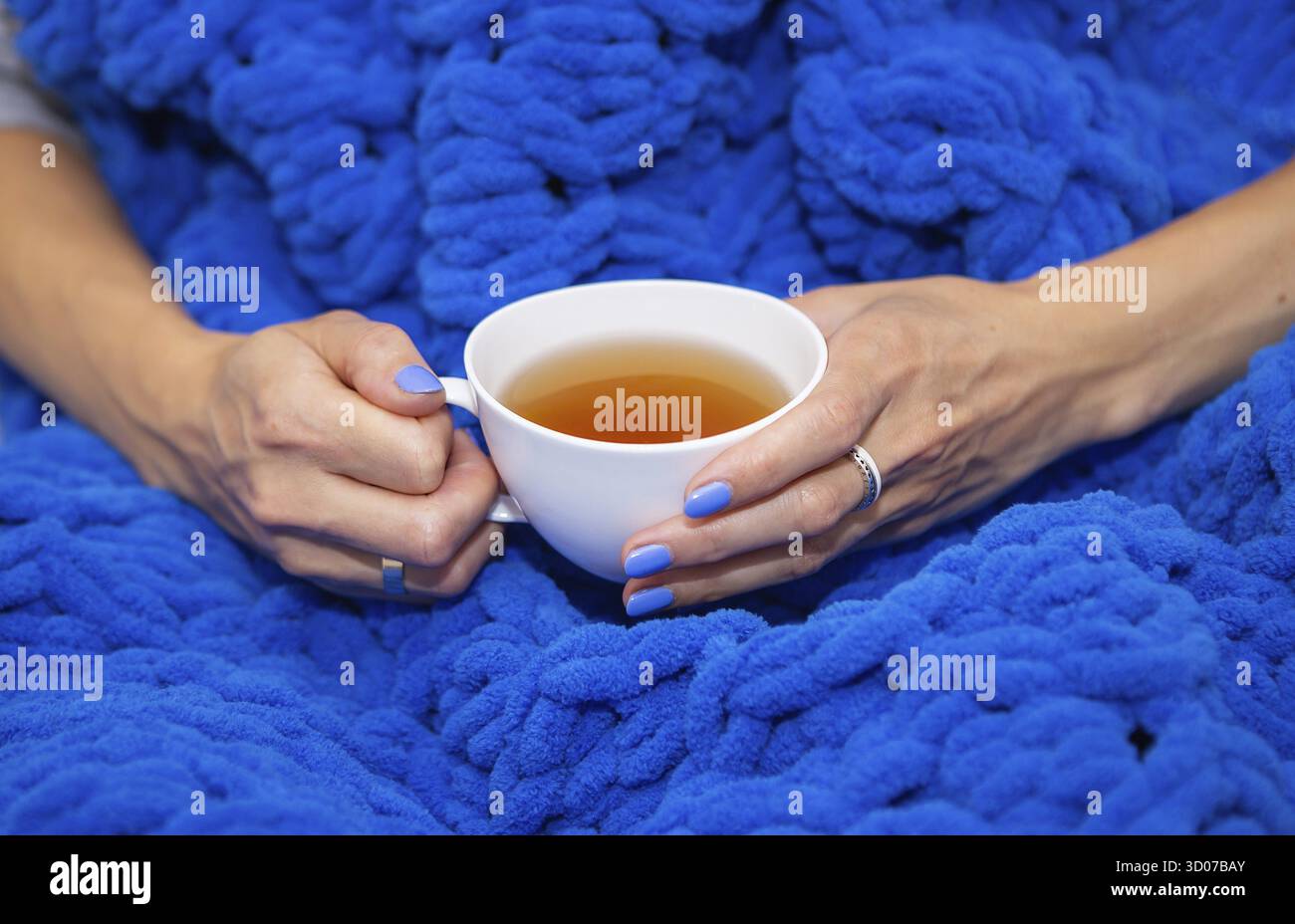 Una ragazza avvolta in una grande coperta maglia a mano di lana merino blu, una coperta calda e molto morbida, una ragazza che tiene una tazza bianca di tè caldo, posto per il testo Foto Stock