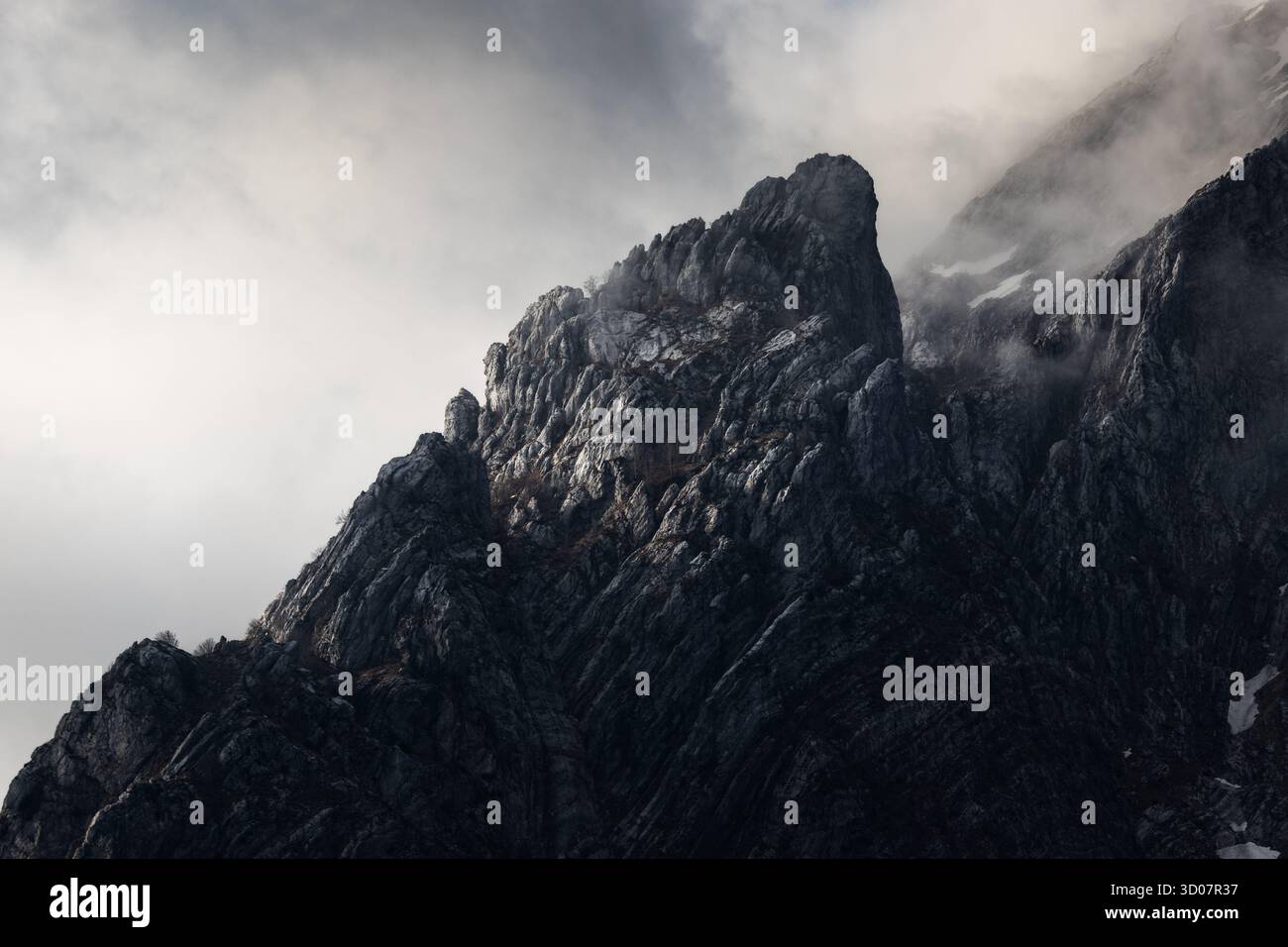 Vista di un'aspra cima di montagna grigia scura che pierga attraverso nuvole vorticose con macchie di neve aggrappate ai suoi lati, Carrara, Toscana, Italia. Foto Stock