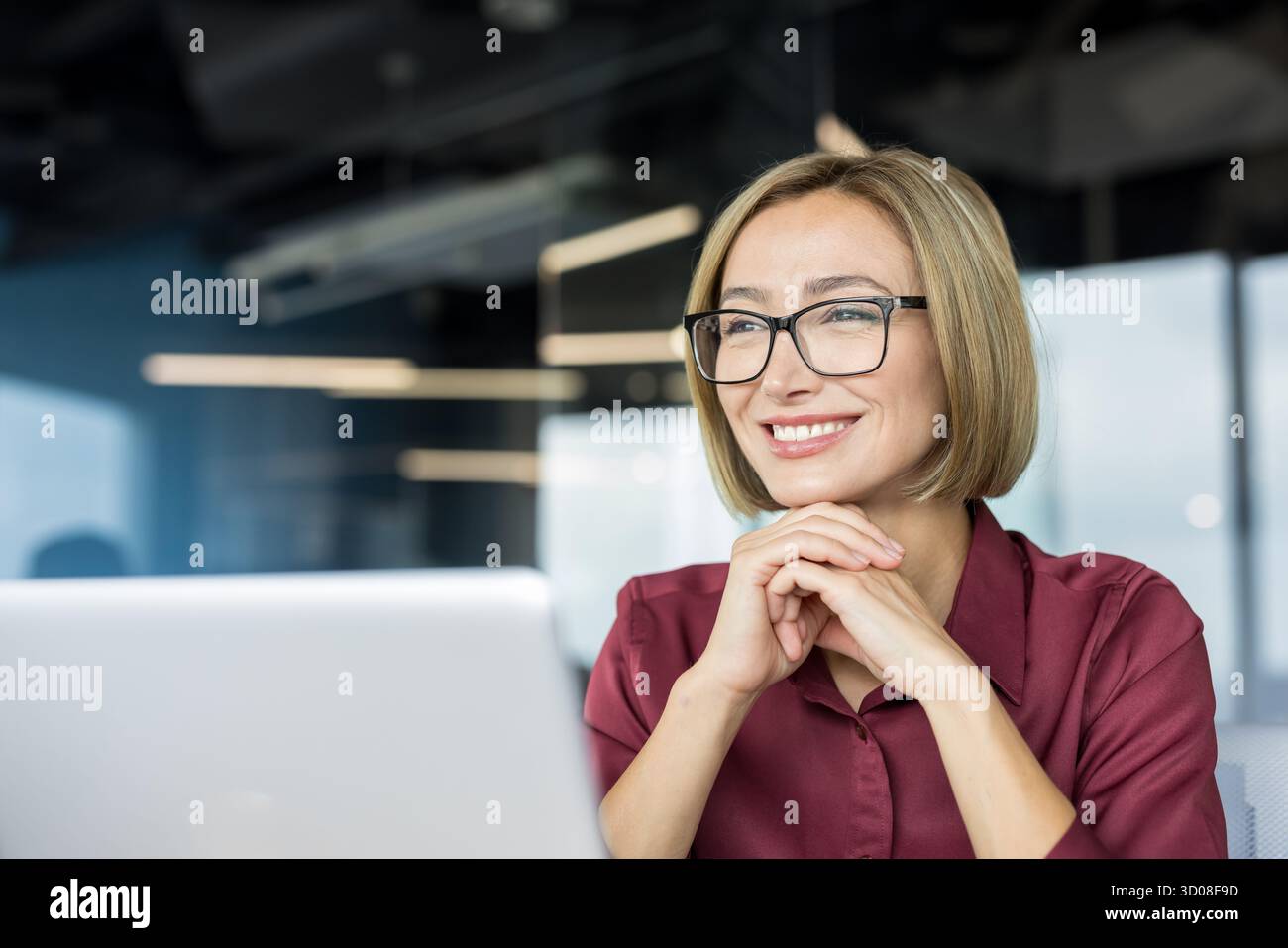 Donna d'affari felice che indossa occhiali da vista e una camicia marrone, sorridendo allegramente mentre lavora su un notebook in un moderno ufficio aziendale Foto Stock