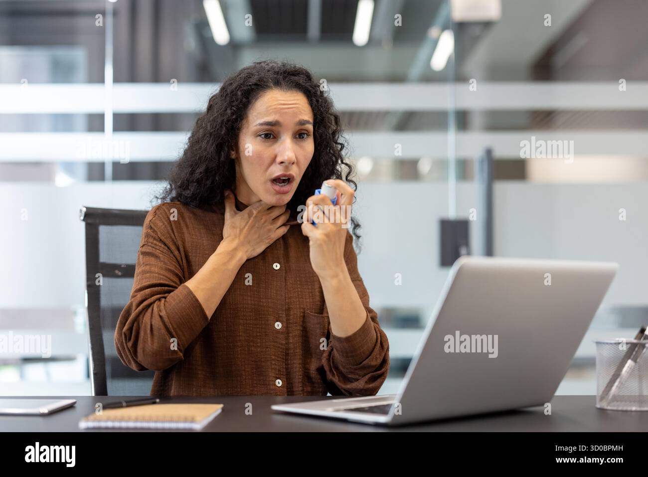 Donna che ha un attacco d'asma al suo portatile in un ufficio moderno, fatica a respirare e usa un inalatore per un sollievo urgente durante una giornata di lavoro stressante Foto Stock
