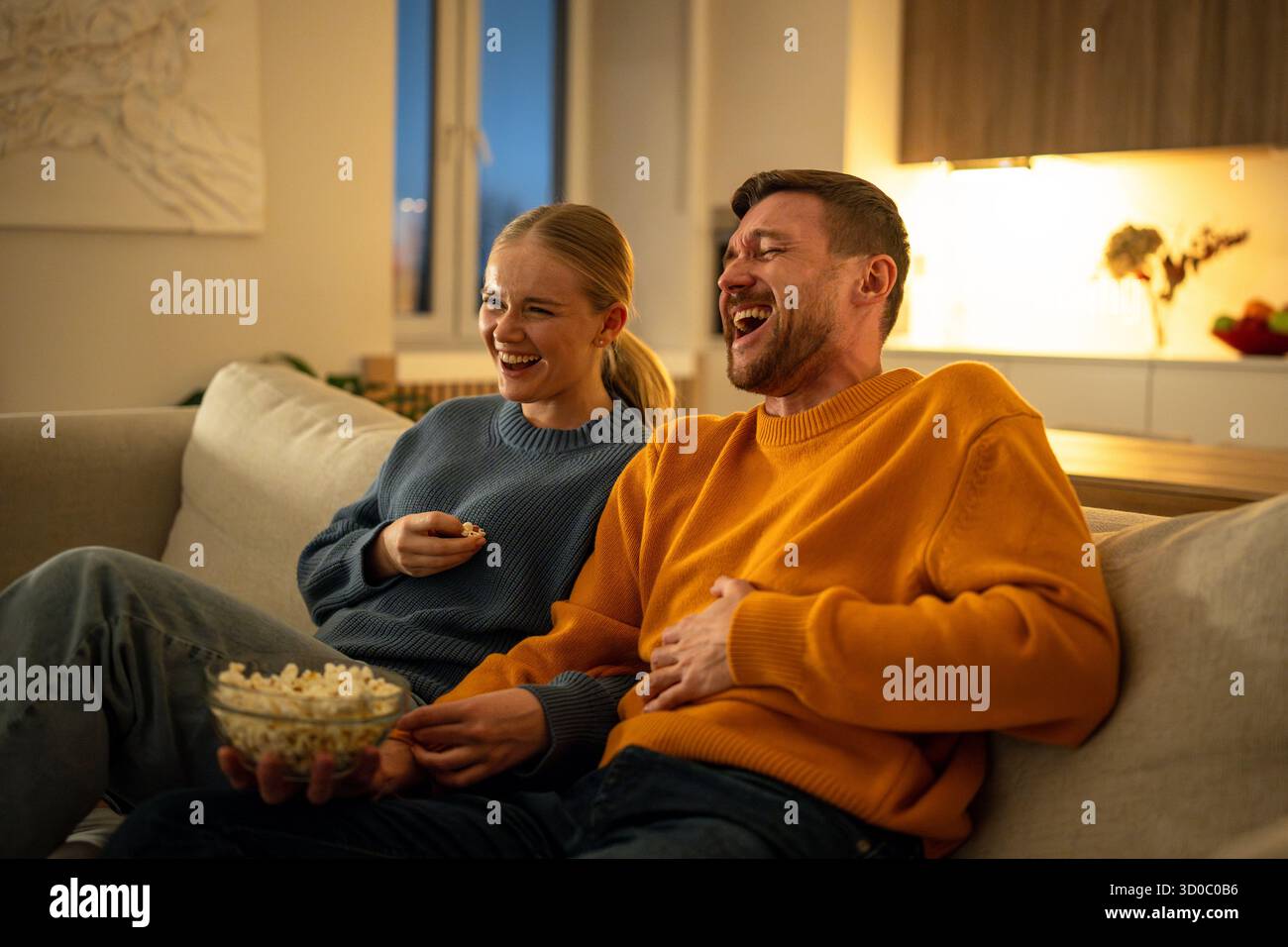 Serata con film in famiglia, giovani coppie che ridono e mangiano popcorn mentre guardano la commedia in una casa accogliente Foto Stock