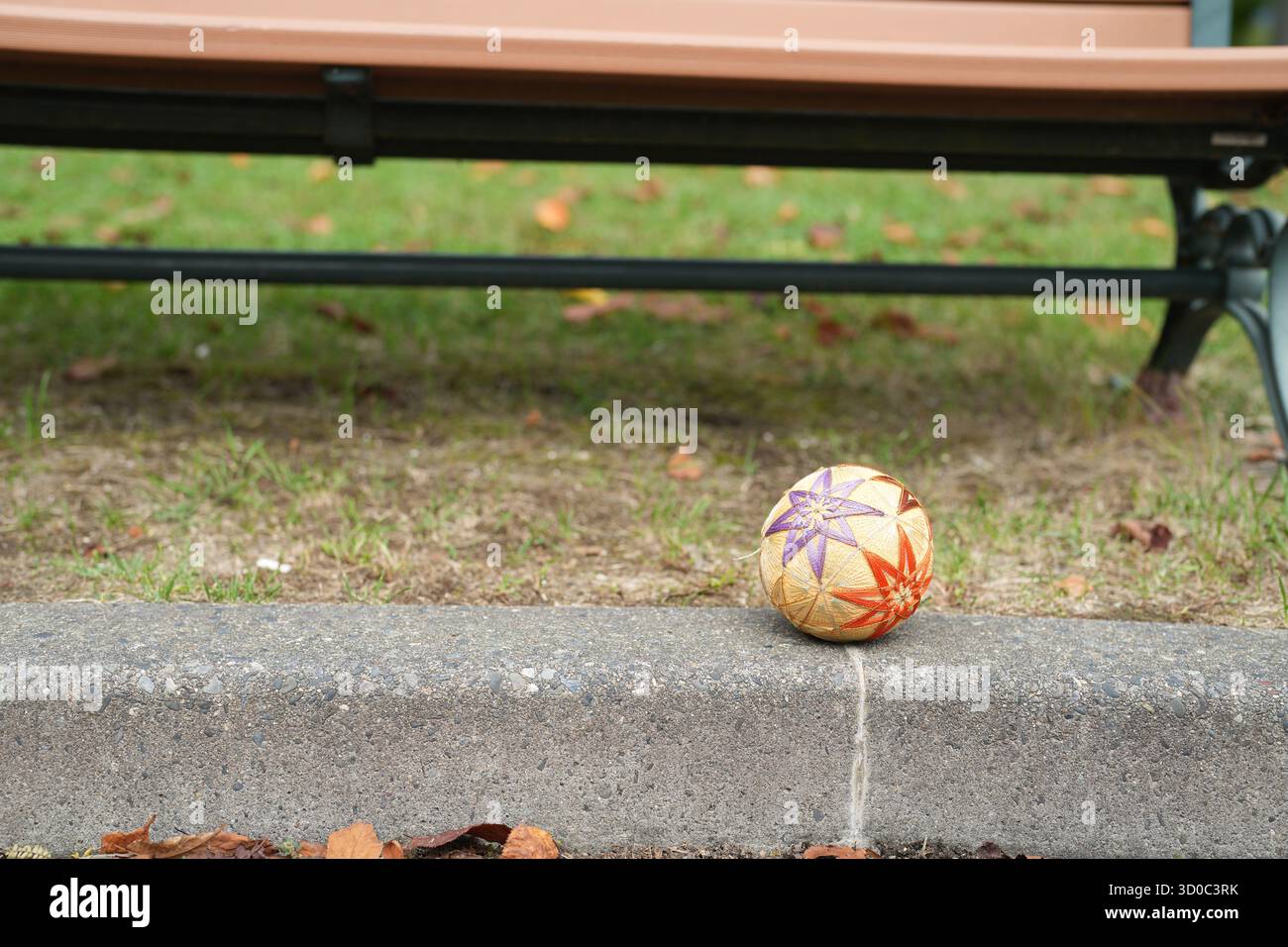 Una piccola palla è seduta su un marciapiede accanto a una panchina. La palla è gialla e viola ed è un oggetto decorativo. La panchina e' vuota e la scena lo e' Foto Stock