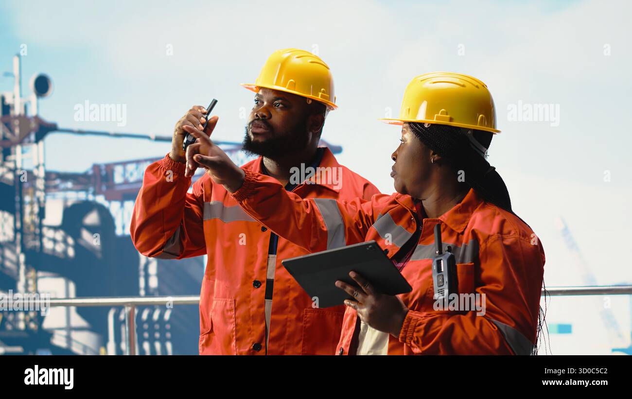 Dipendenti della piattaforma offshore che supervisionano le operazioni del deck con il dispositivo, parlando al walkie talkie. Ingegneri afroamericani che effettuano manutenzione su attrezzature per trivellazioni, trasmettendo messaggi radio Foto Stock