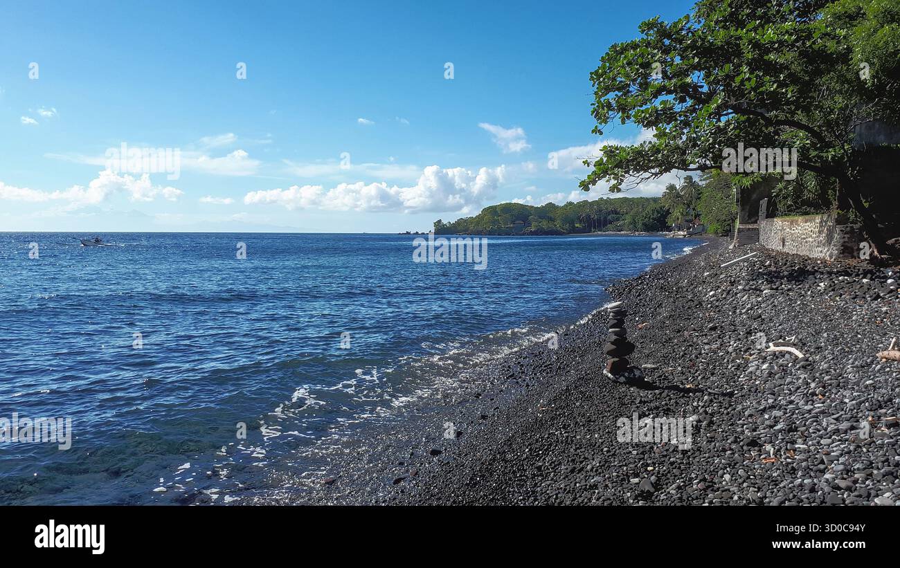 Costa lungo la costa nord-est di Bali con una spiaggia di ciottoli vulcanici scuri e una torre di ciottoli accuratamente impilata, che mette in risalto le texture naturali Foto Stock