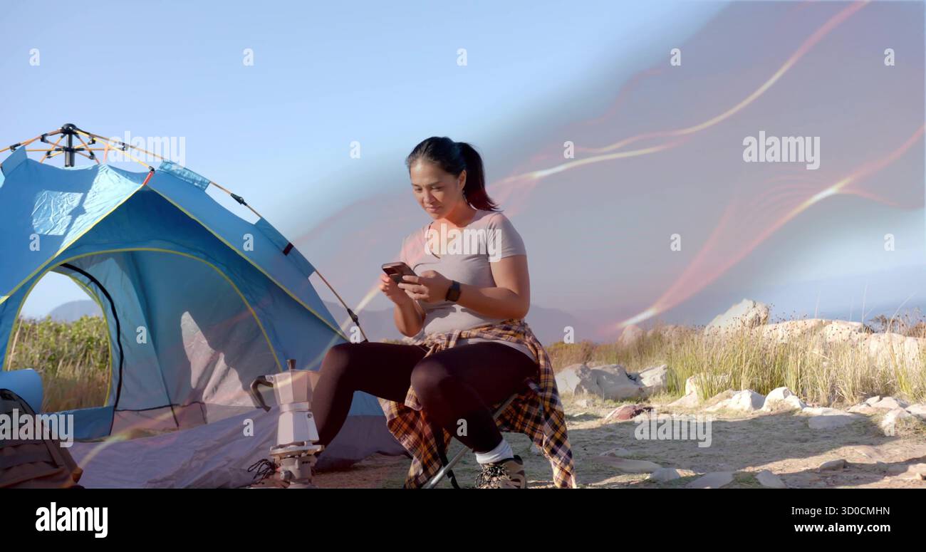 Una donna con smartphone indossa l'attrezzatura da trekking seduta sullo sgabello in un campeggio rurale con tenda e treppiede Foto Stock