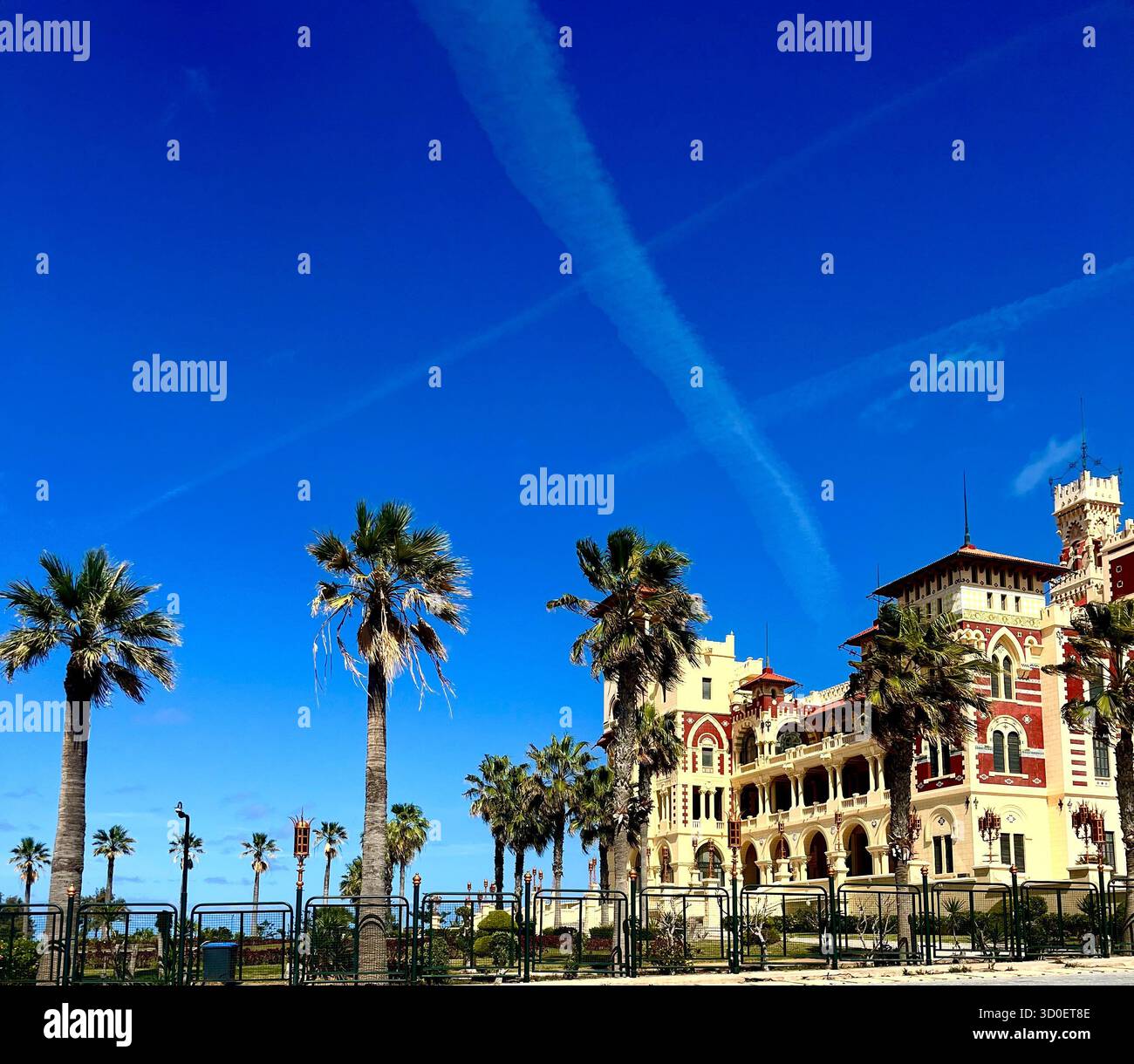 Storico edificio in stile mediterraneo con palme sotto un cielo azzurro luminoso e contrafforti di aerei, catturato in una giornata di sole sulla costa. - Immagine stock catturata con smartphone