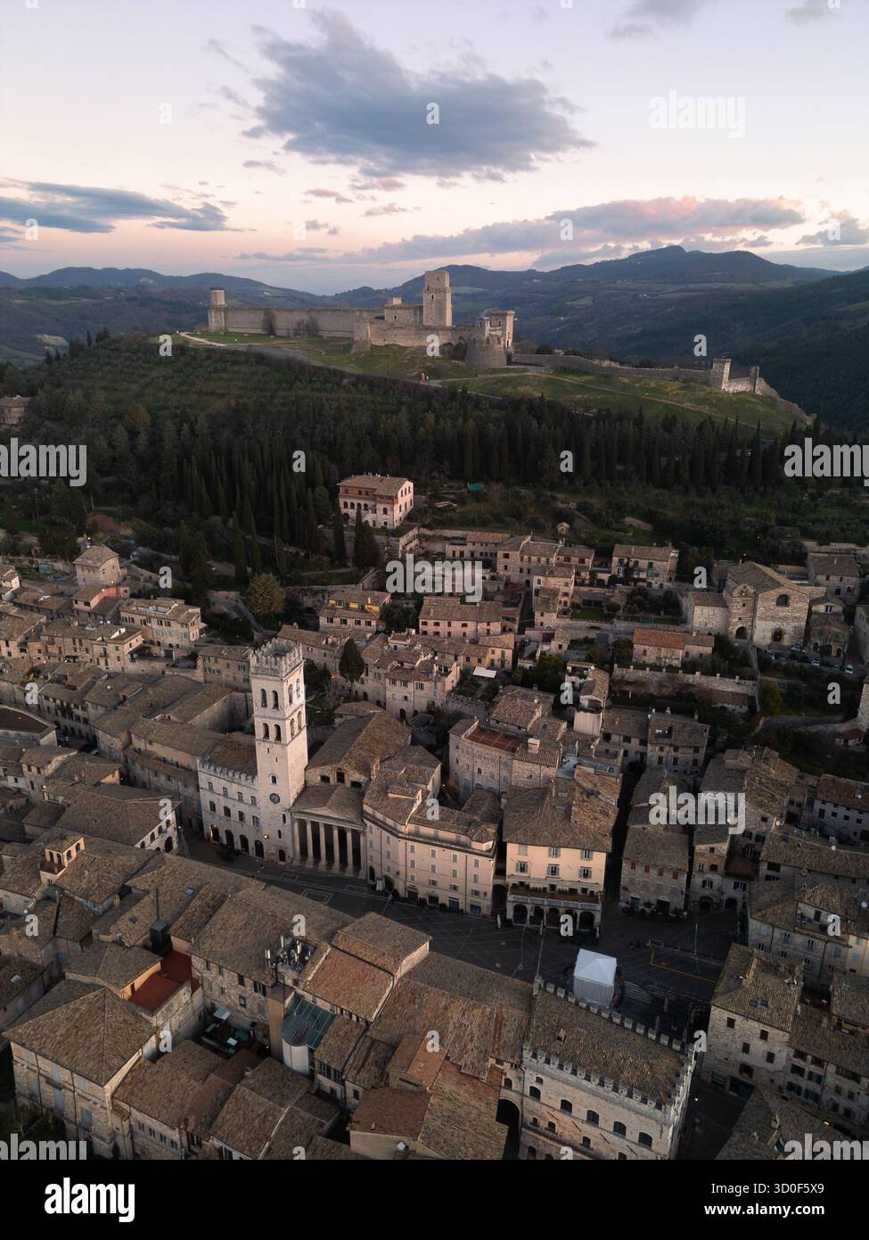Veduta aerea della Basilica Papale di San Francesco d'Assisi che si erge maestosamente in cima a una collina verdeggiante, mentre la pittoresca cittadina si dispiega sotto in un arazzo di tetti in terracotta, Assisi, Umbria, Italia. Foto Stock