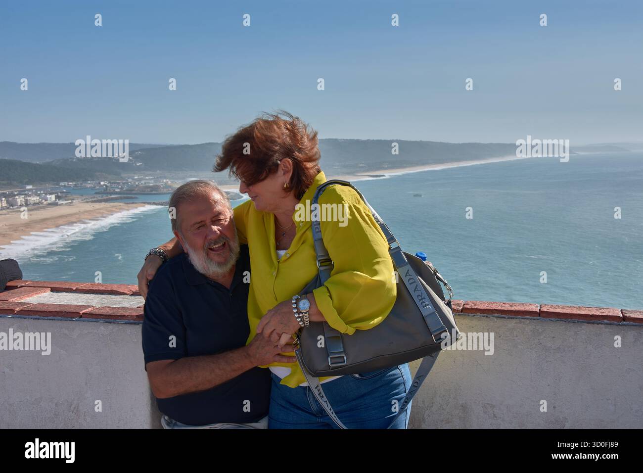 Una coppia più anziana si pone teneramente abbracciando un punto panoramico a Nazare, in Portogallo, con l'Oceano Atlantico e la costa sullo sfondo. Foto Stock