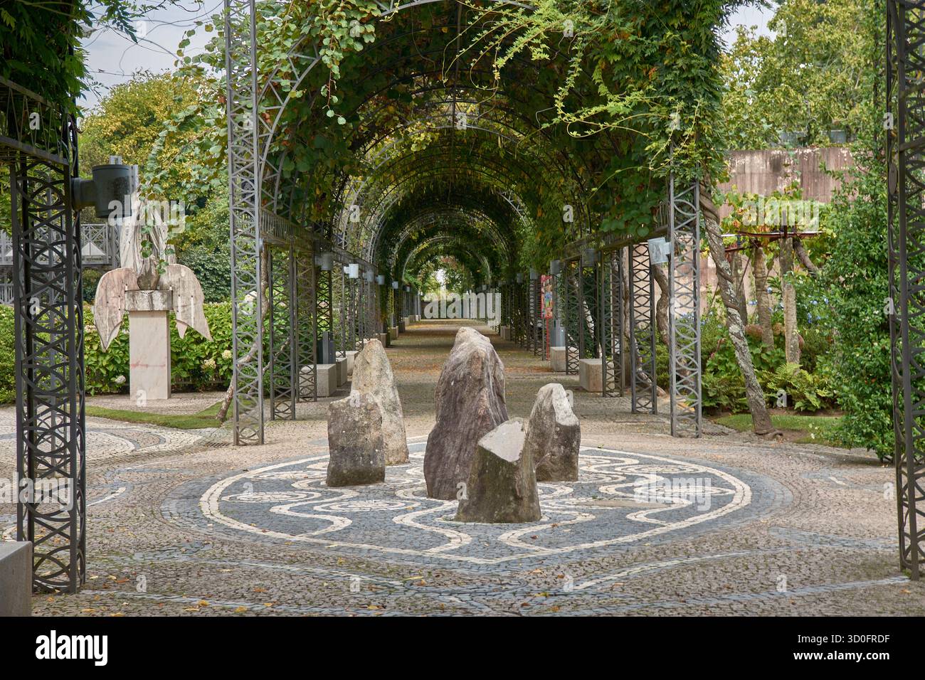 Un tunnel di piante costituito da archi metallici ricoperti di fogliame guida il visitatore lungo un percorso a mosaico in un lussureggiante giardino. In primo piano, pietre verticali Foto Stock