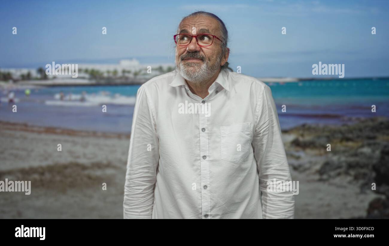 Uomo con occhiali rossi sorridente e in piedi sulla spiaggia vicino agli edifici del resort e alla spiaggia rocciosa, camicia con bottoni, mani rilassate ai lati; serenità. Foto Stock