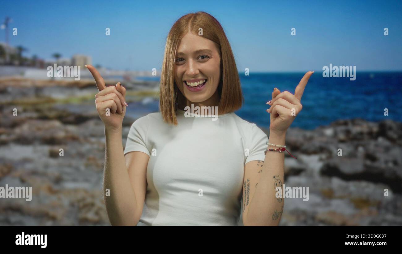 La giovane donna bionda punta le dita indice verso l'alto sulla costa rocciosa del mare soleggiato; ottimismo entusiasmo eccitazione gioia. Foto Stock