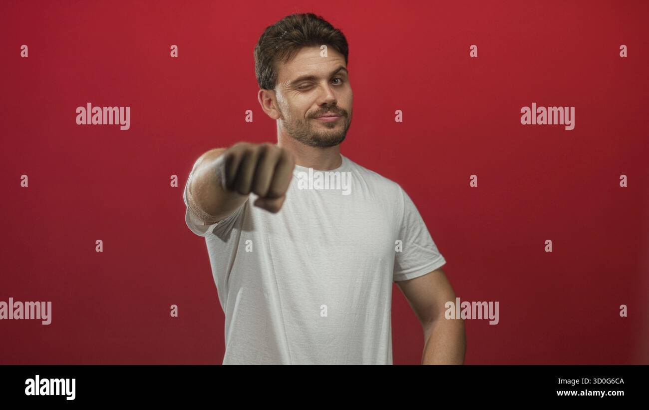Giovane ispanico in t shirt bianca che si estende il pugno verso la fotocamera mentre si fa l'occhiolino in studio rosso; fascino di fiducia. Foto Stock