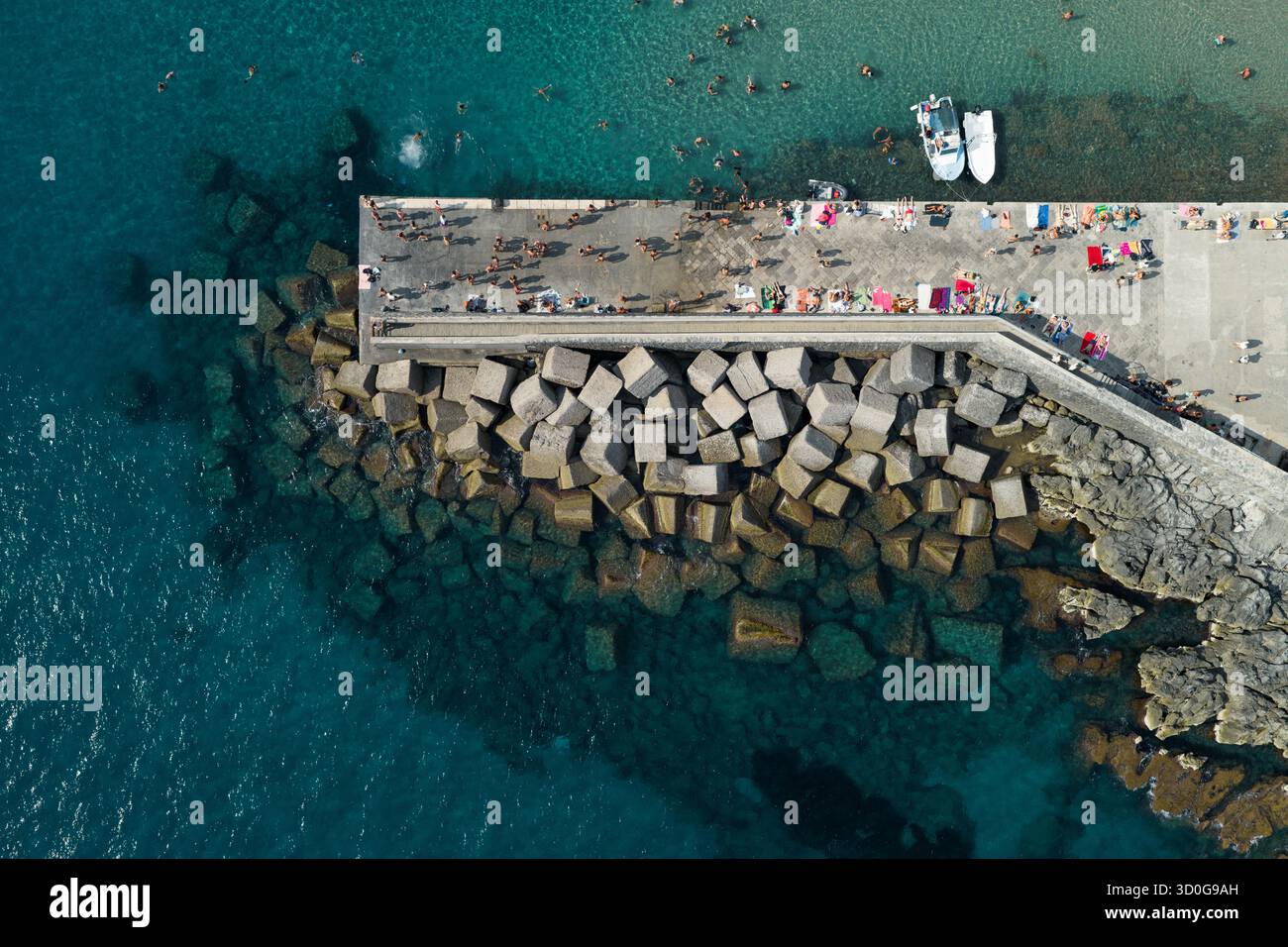 Vista aerea dei bagnanti che si crogiolano sul molo, contrastando con le acque turchesi e la frastagliata frangiflutti, Cefalù, Sicilia, Italia. Foto Stock
