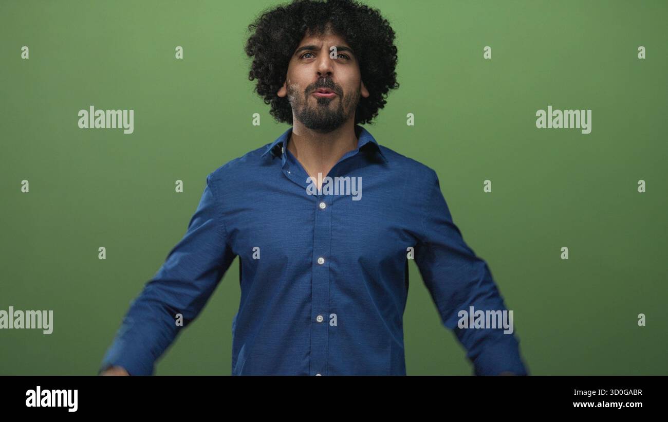 Uomo con capelli ricci che indossa una camicia blu e un orologio da polso, che alza i pugni e sorride verso la fotocamera in uno studio verde; emozione. Foto Stock