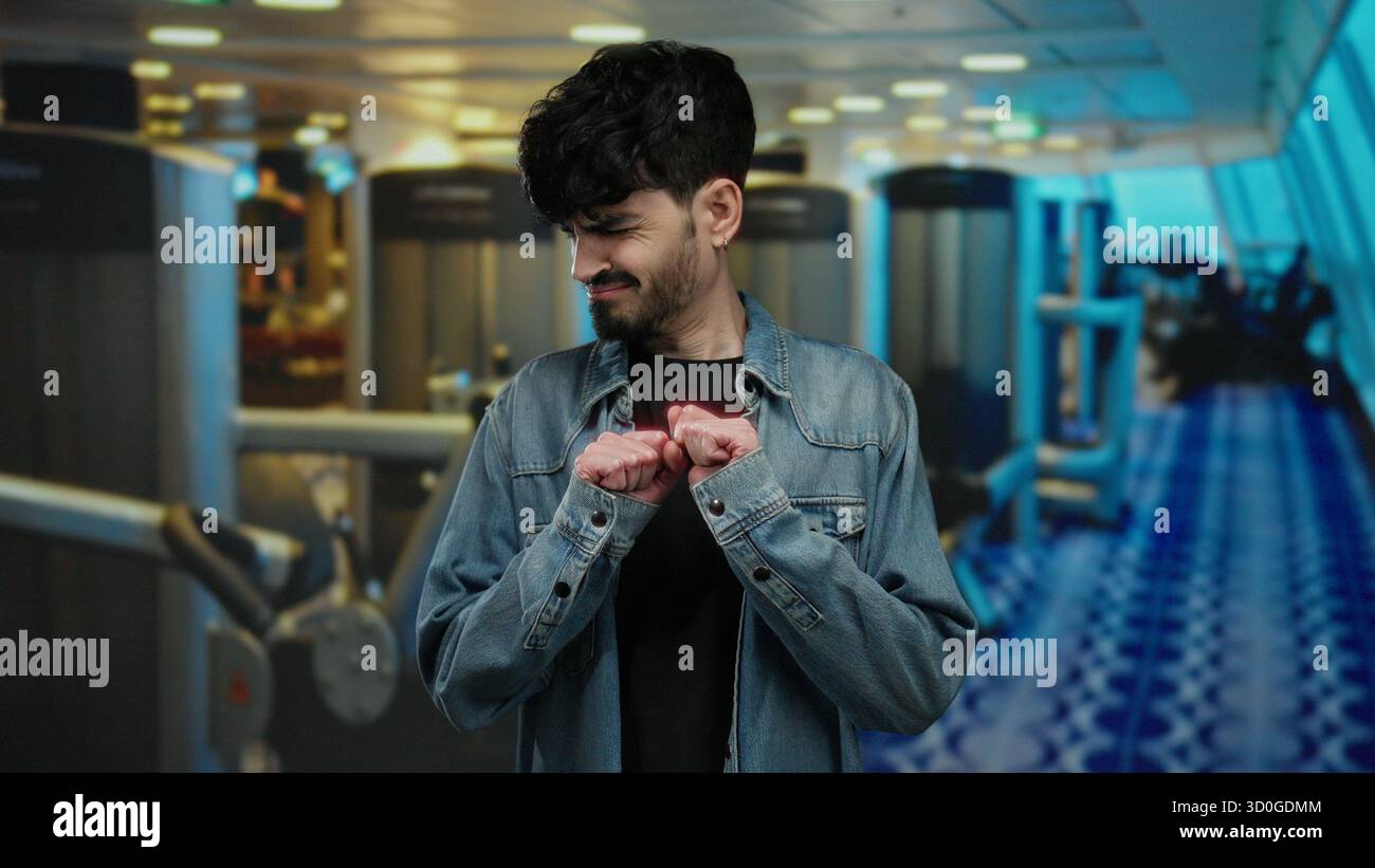 Giovane uomo che esprime disprezzo in palestra, indossa una giacca in denim sullo sfondo di moderne attrezzature fitness. Foto Stock
