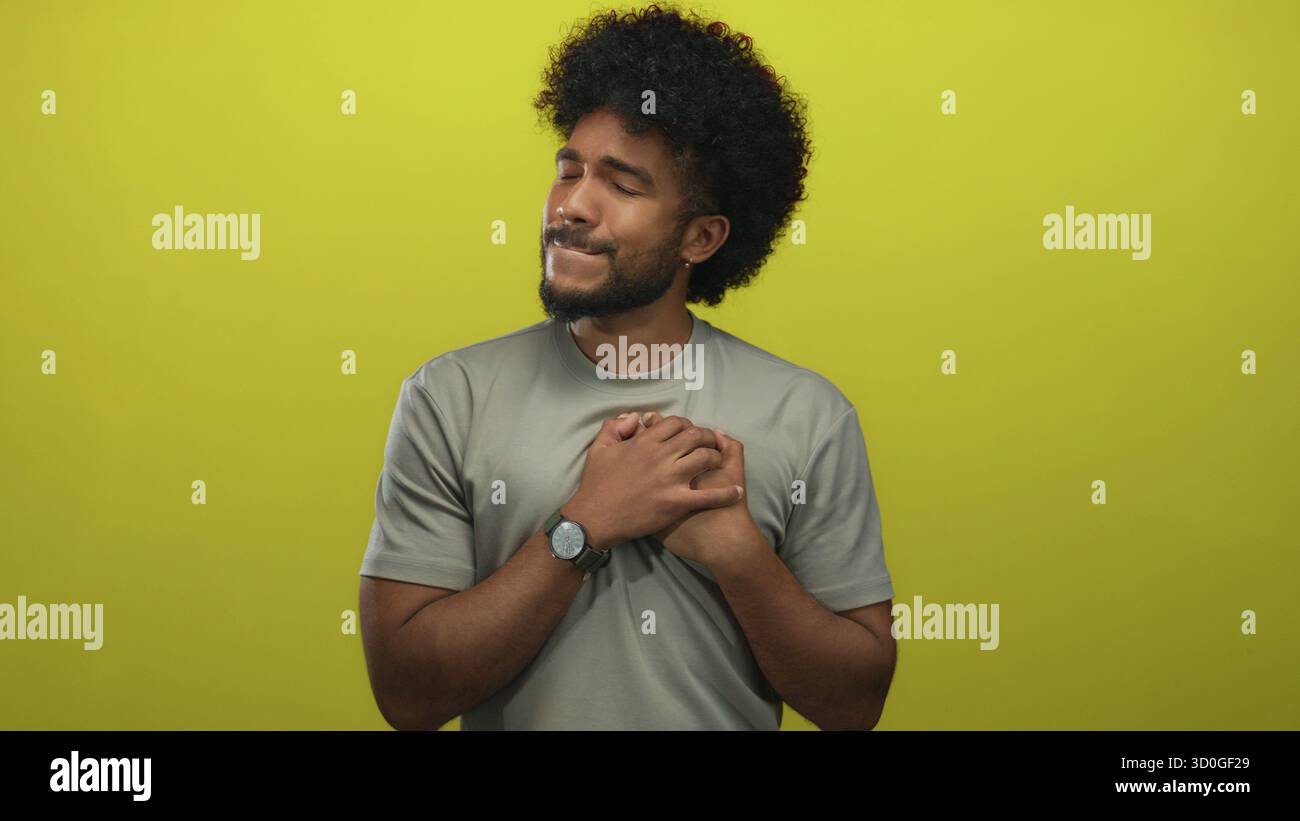 Uomo che tiene il petto davanti al muro giallo con un'espressione dolorosa, indossa una camicia grigia, capelli ricci e guarda, trasmettendo emozione e solitudine. Foto Stock