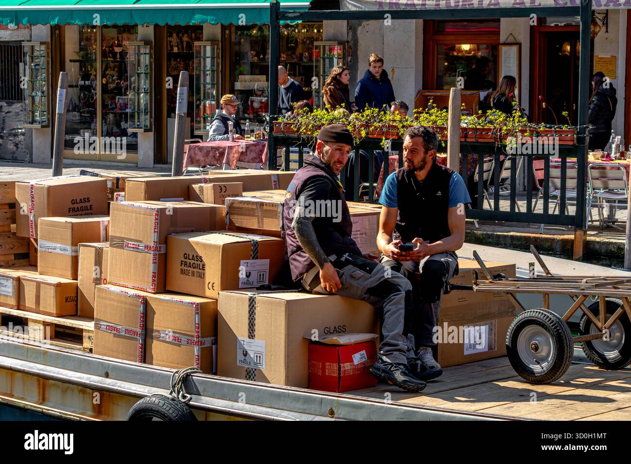 Due uomini in conversazione seduti su scatole di cartone su una chiatta merci che fa consegne attraverso un canale a Venezia, in Italia Foto Stock
