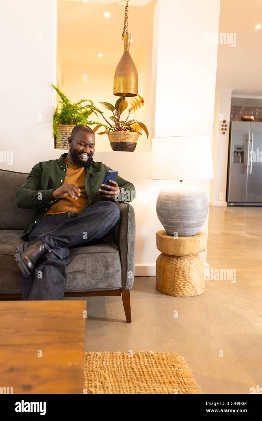 Uomo afro-americano seduto sul divano con uno smartphone sotto la lampada a sospensione di casa, spazio fotocopie Foto Stock