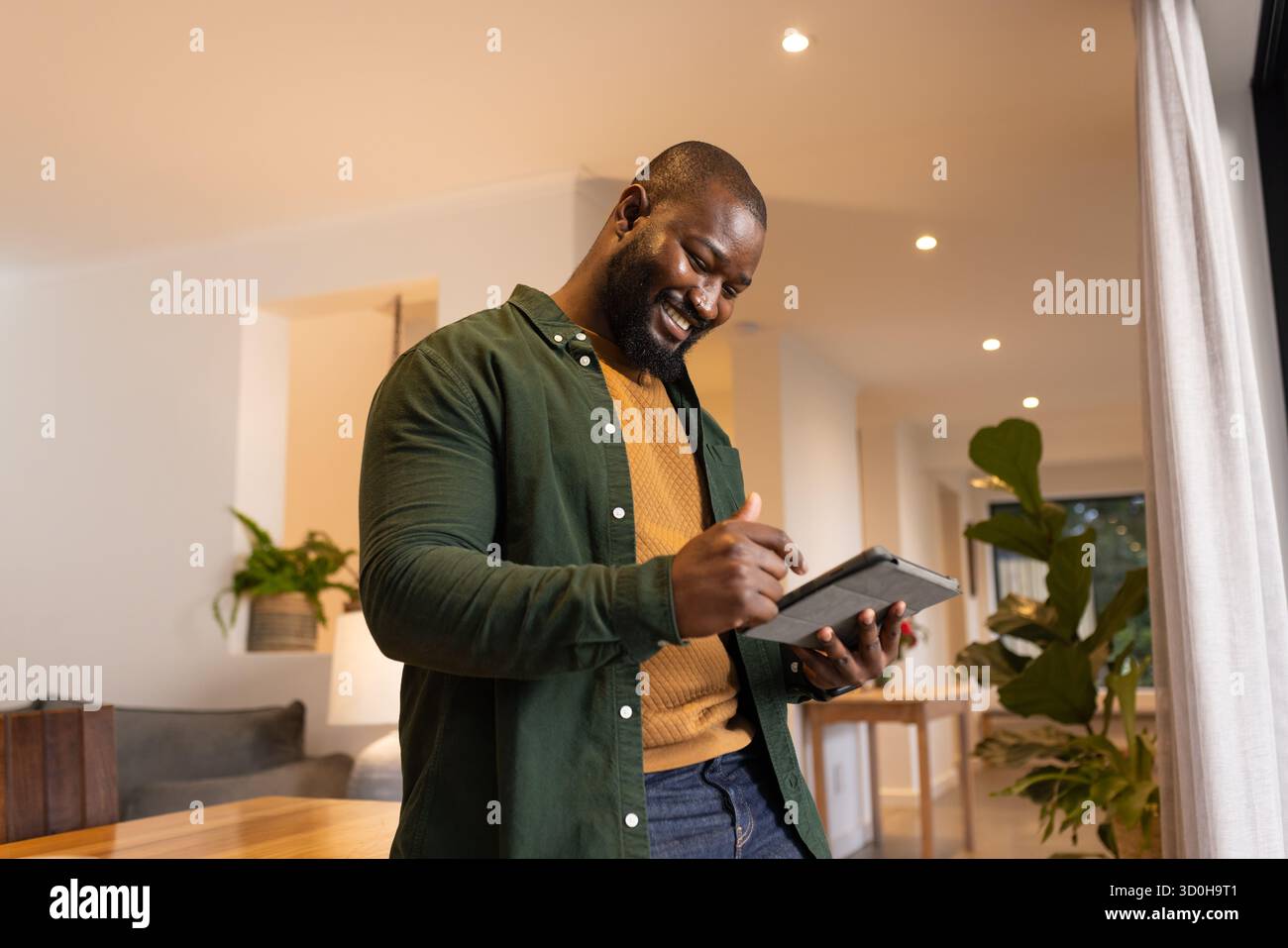 Uomo afroamericano in piedi in salotto a scrivere su tablet con stilo e maglione senape Foto Stock
