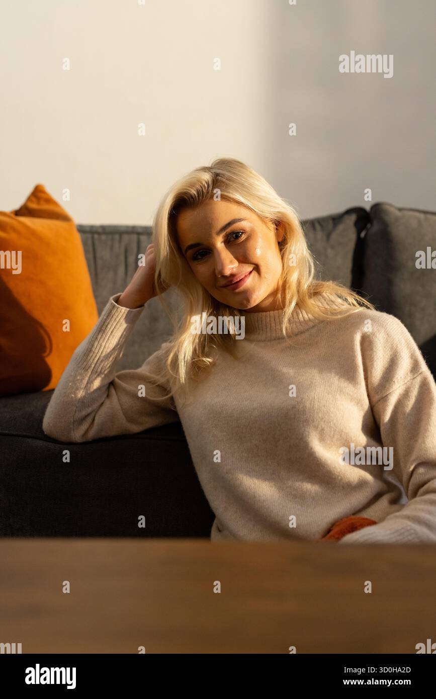 Donna seduta su un divano grigio scuro, appoggiata al gomito, sorridente in una stanza illuminata dal sole con cuscino arancione Foto Stock