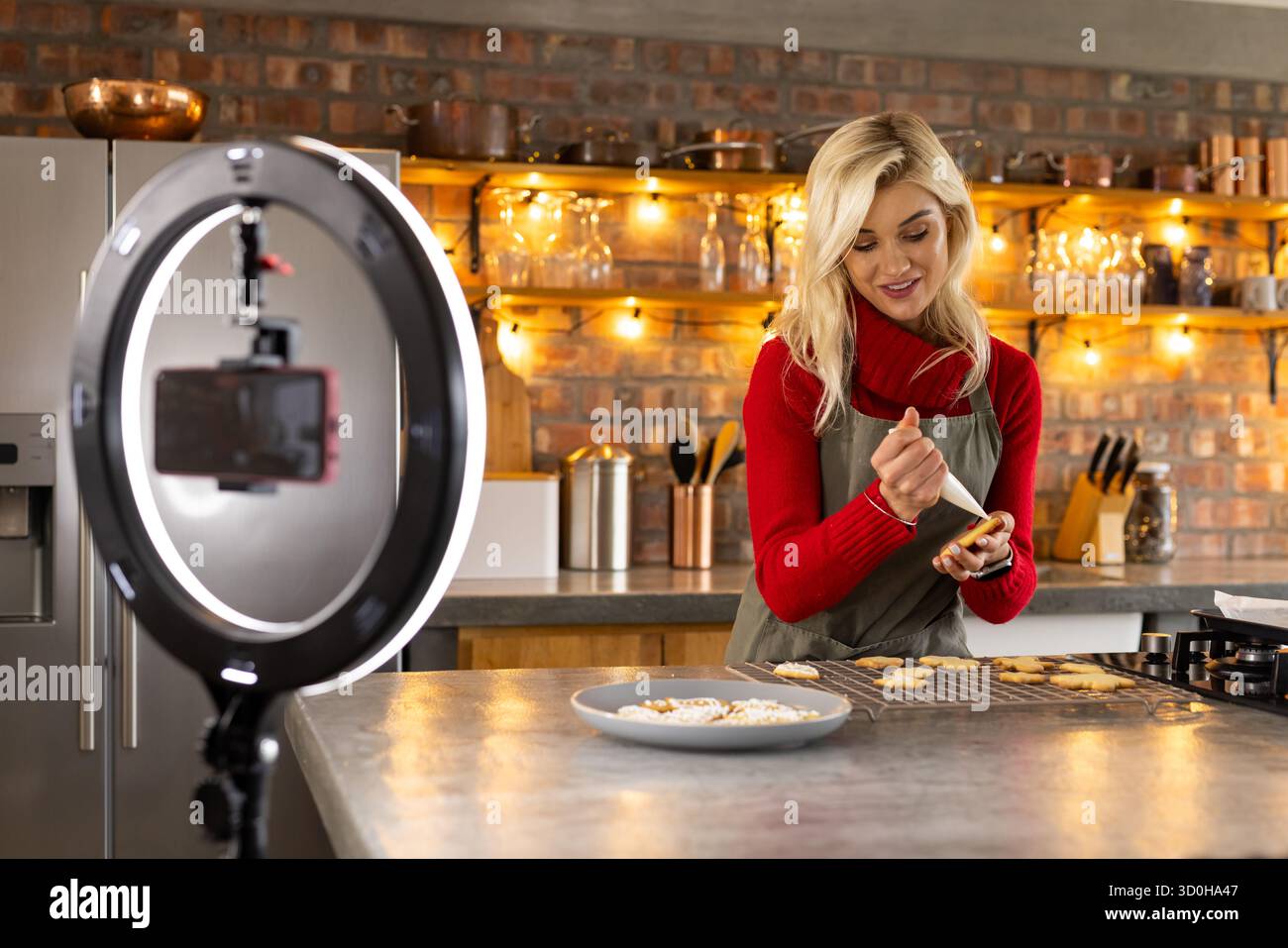 panettiera che decora biscotti su scaffale in cucina indossando grembiule verde sotto il semaforo anulare con il telefono Foto Stock