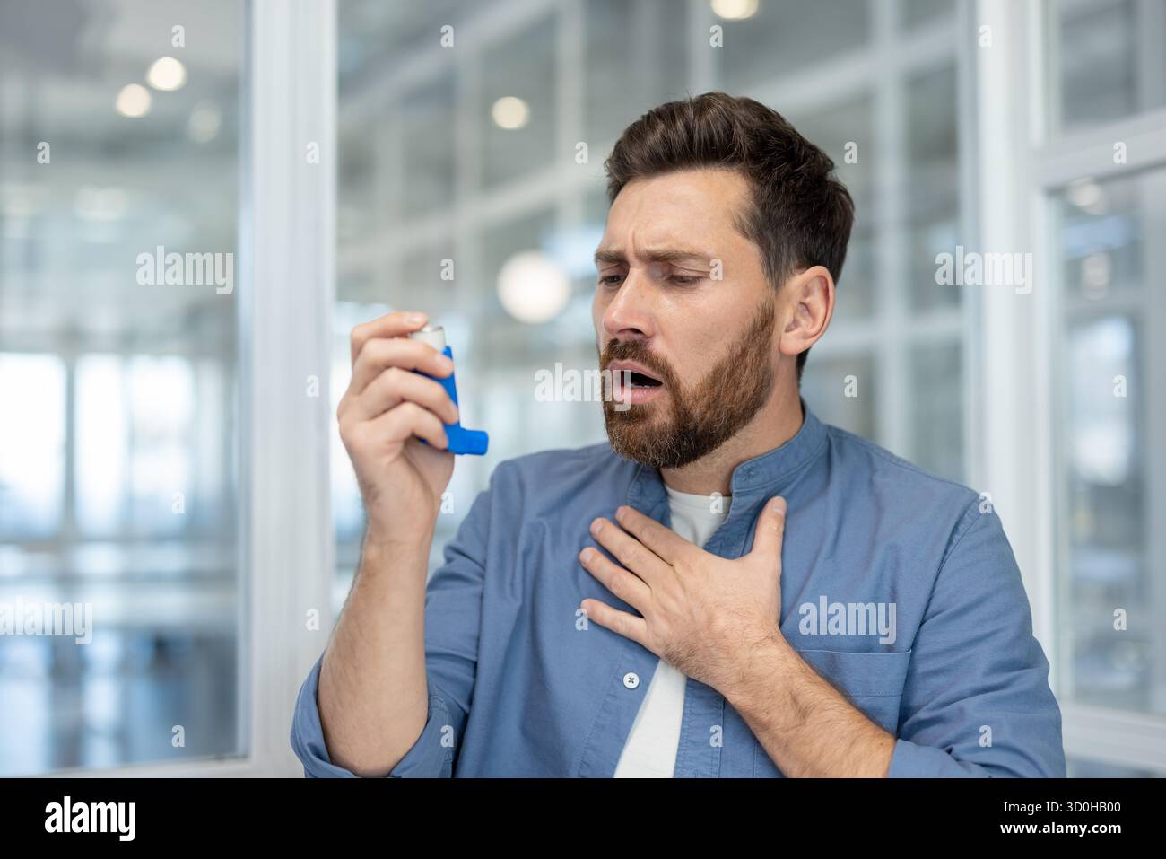 Uomo barbuto che soffre di un attacco di asma acuto, stringendo il petto e tenendo in mano un inalatore, lottando per respirare e apparendo ansioso e senza fiato in interni, primo piano Foto Stock