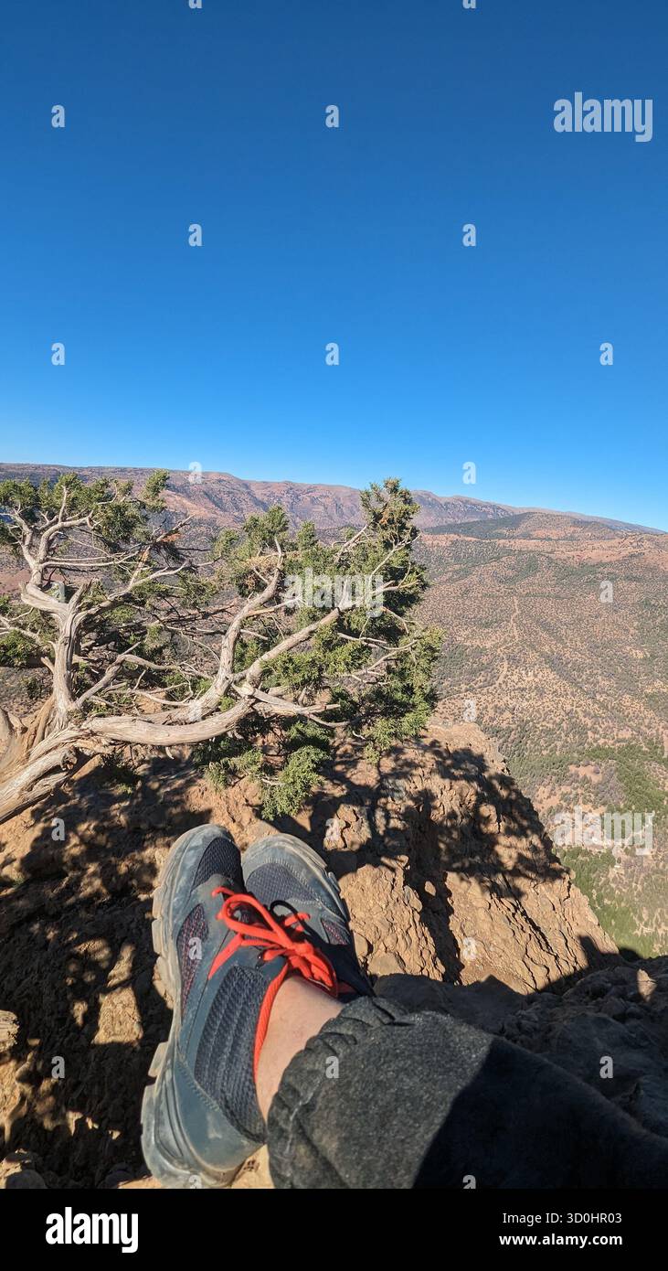 Punto di vista degli scarponi da trekking sul bordo di una scogliera che si affaccia su un vasto paesaggio secco di montagna sotto un cielo blu Foto Stock