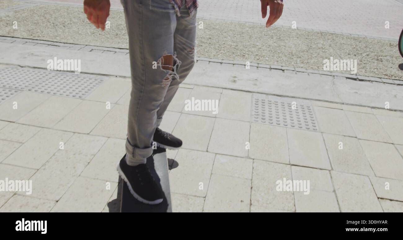 Skateboarder maschile che indossa jeans strappati e sneakers sul marciapiede con tegole e skateboard Foto Stock