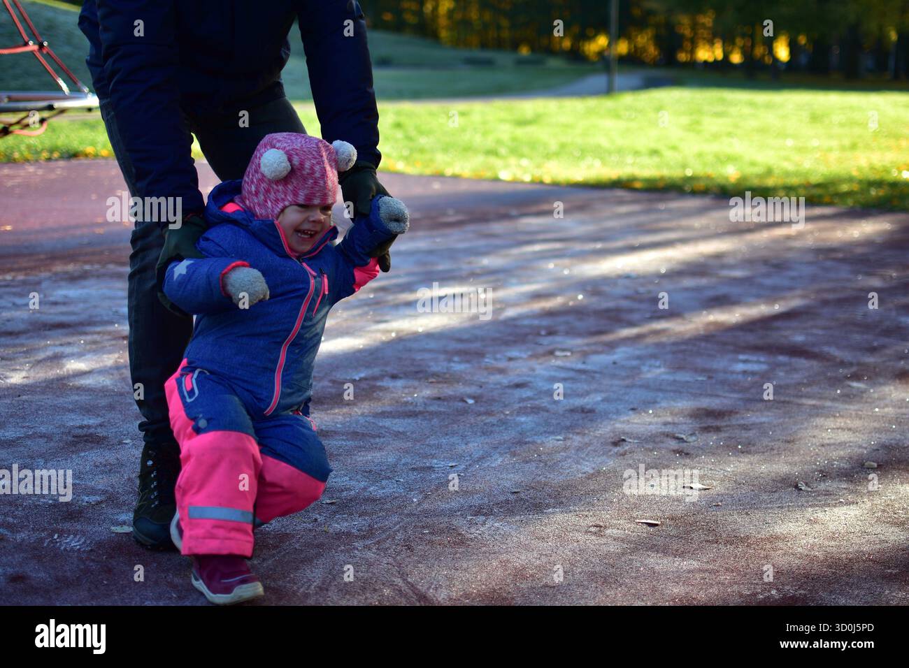 Una bambina che ride con le tute invernali impara a camminare sul parco giochi ghiacciato con i genitori aiuta nella soleggiata mattinata autunnale. Un momento di gioia per tutta la famiglia all'aperto Foto Stock