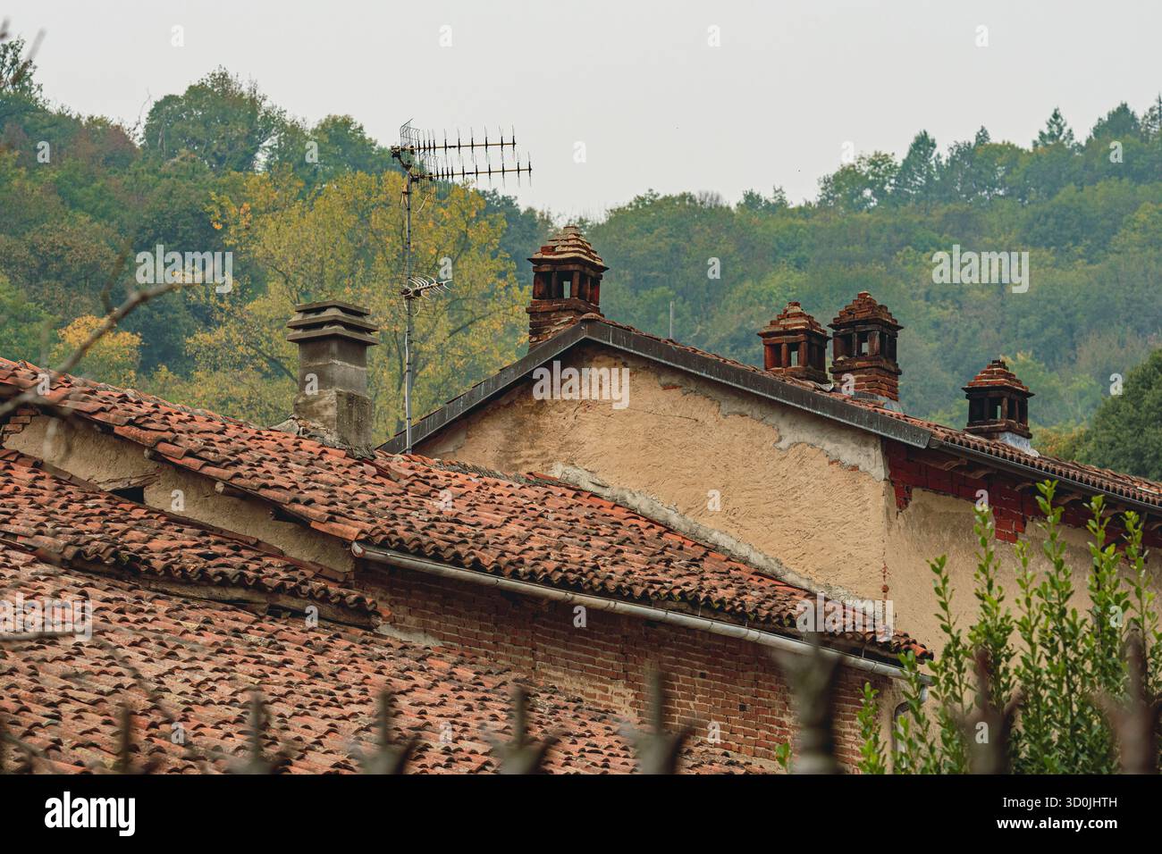 Tradizionali tetti italiani a Monteu da po, in Piemonte, con piastrelle rustiche in terracotta, vecchi camini e un'antenna sullo sfondo di lussureggianti Foto Stock