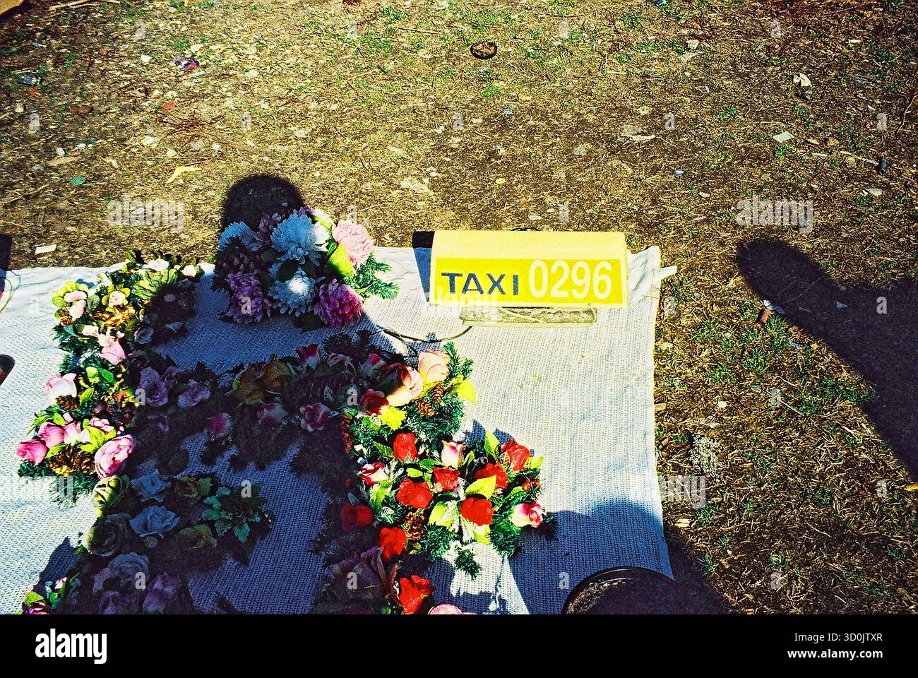 L'ombra del fotografo cade su una stalla per terra che vende fiori di plastica e un cartello taxi, catturato in uno stile analogico incrociato. Foto Stock