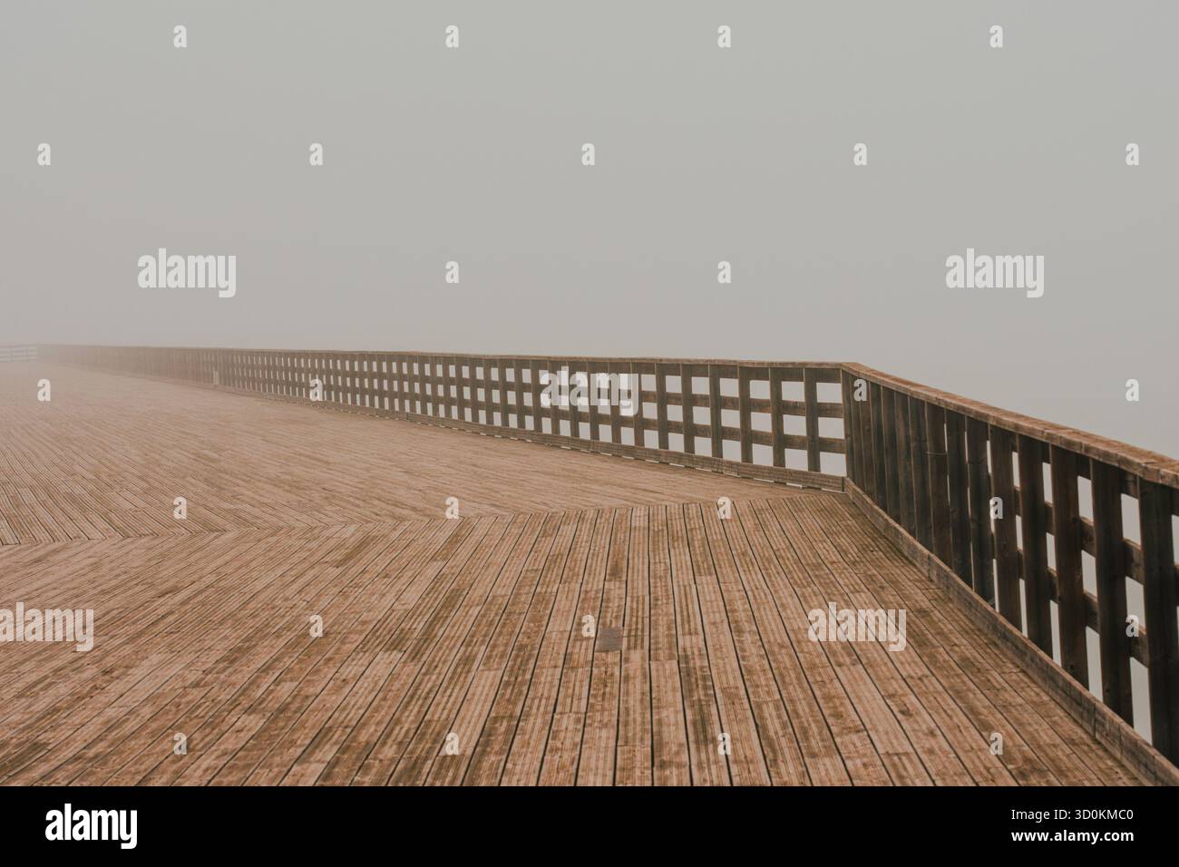 Passerella in legno con ringhiere che sbiadono in fitta nebbia. Le linee geometriche e la luce soffusa creano un'atmosfera tranquilla e misteriosa. Foto Stock