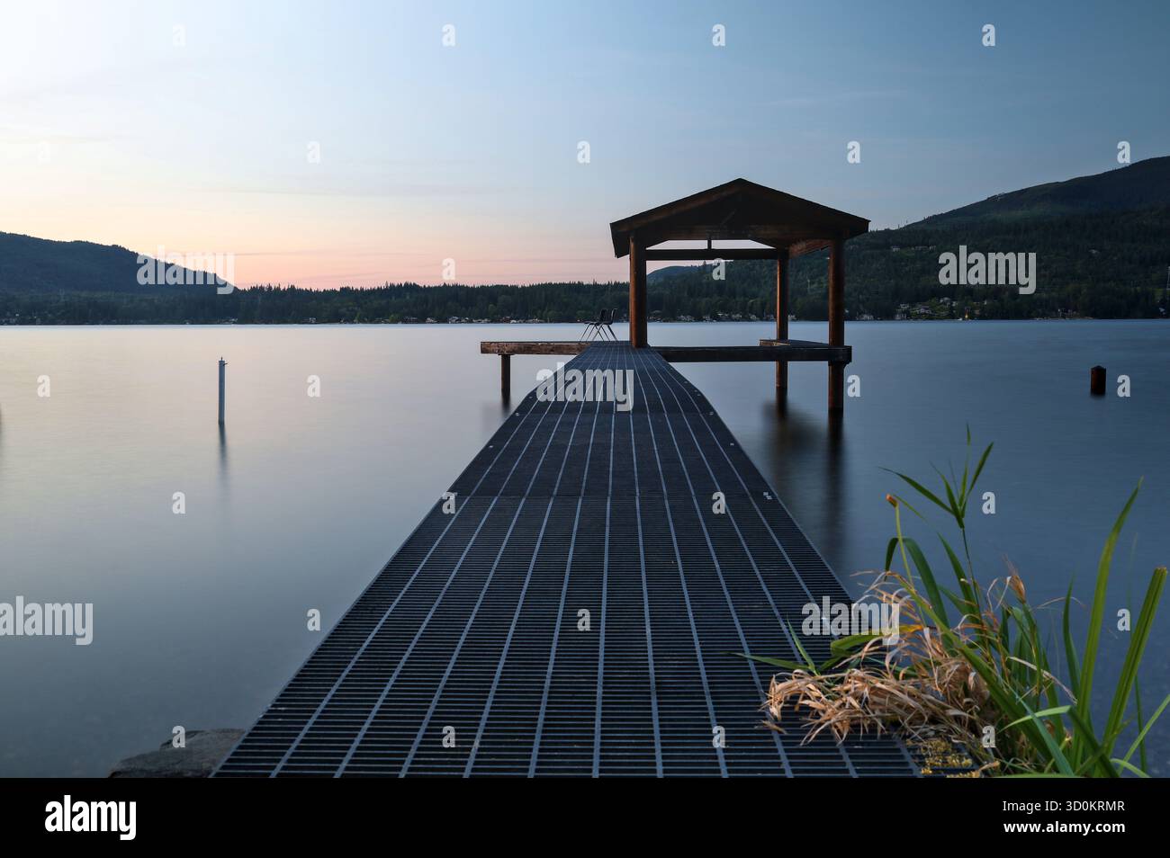 Serena scena sul lago al crepuscolo con un molo di metallo che conduce ad un molo coperto. L'acqua calma riflette il morbido cielo pastello, con le montagne. Foto Stock