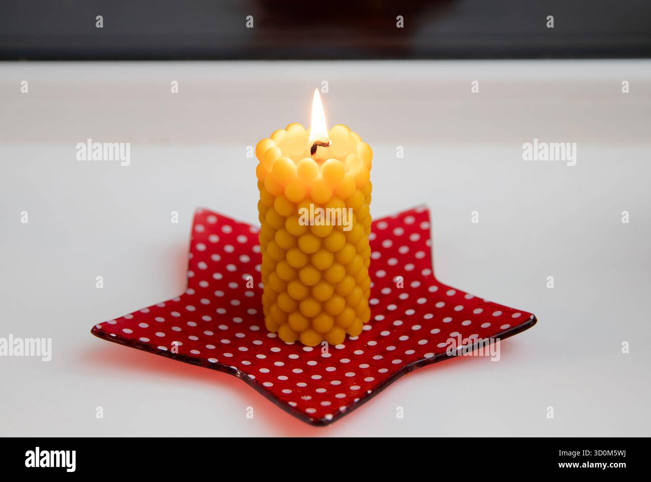 Una candela in cera d'api con motivo spumante, realizzata in una muffa in silicone fatta a mano, brucia su un davanzale a casa. Un piatto rosso a forma di stella. Foto Stock